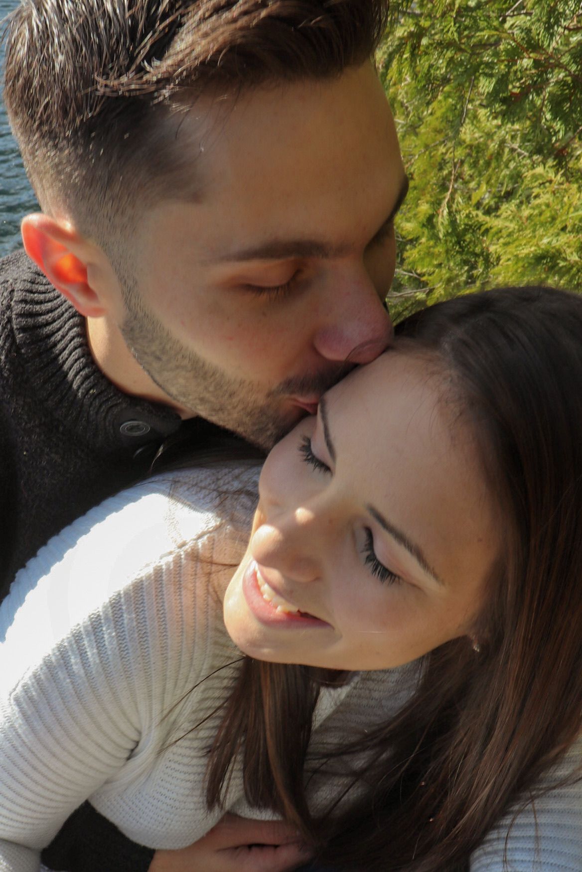 A man tenderly kisses a smiling woman on the cheek outdoors with green foliage in the background.