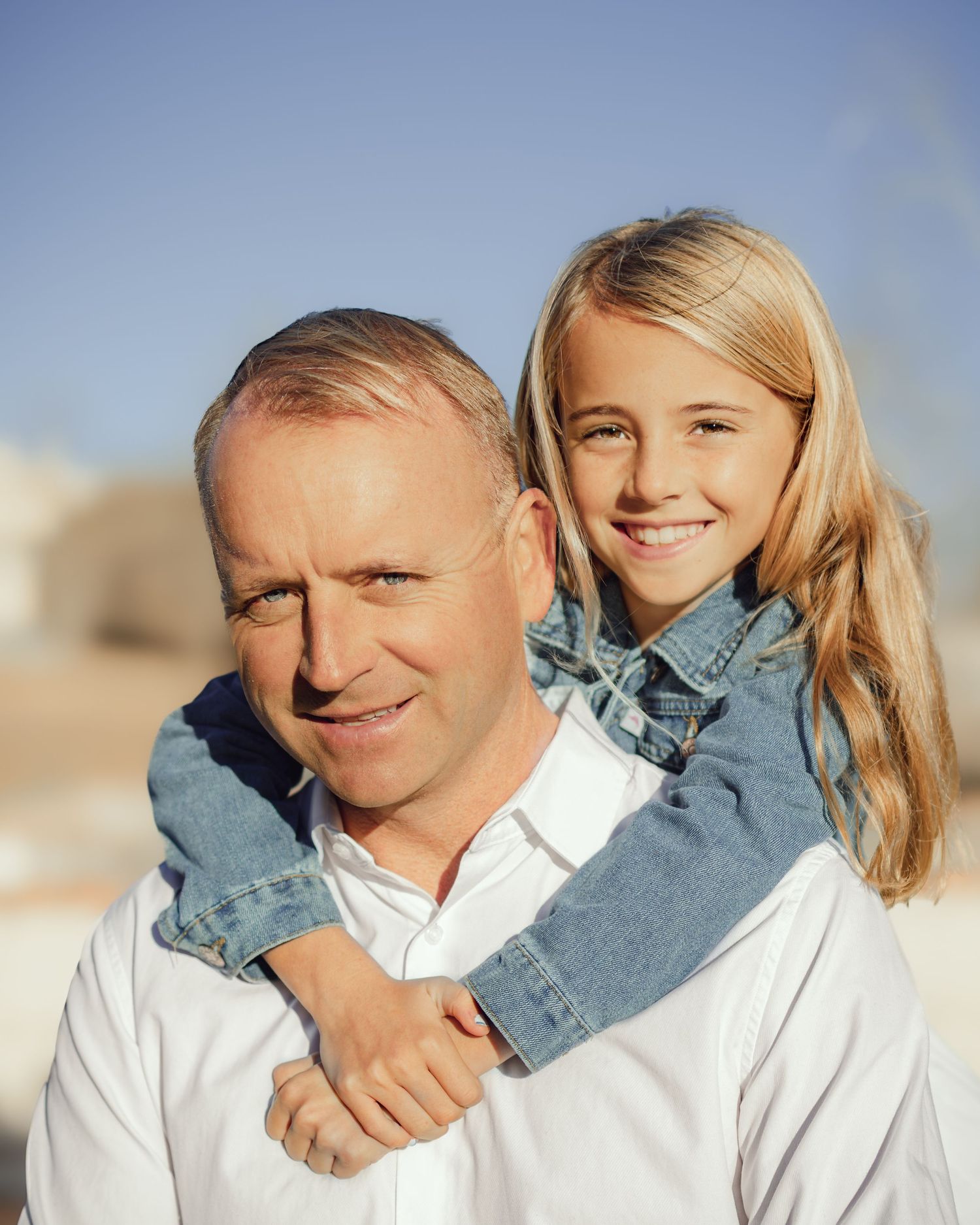 A parent and child share a joyful piggyback moment outdoors against a blue sky background.