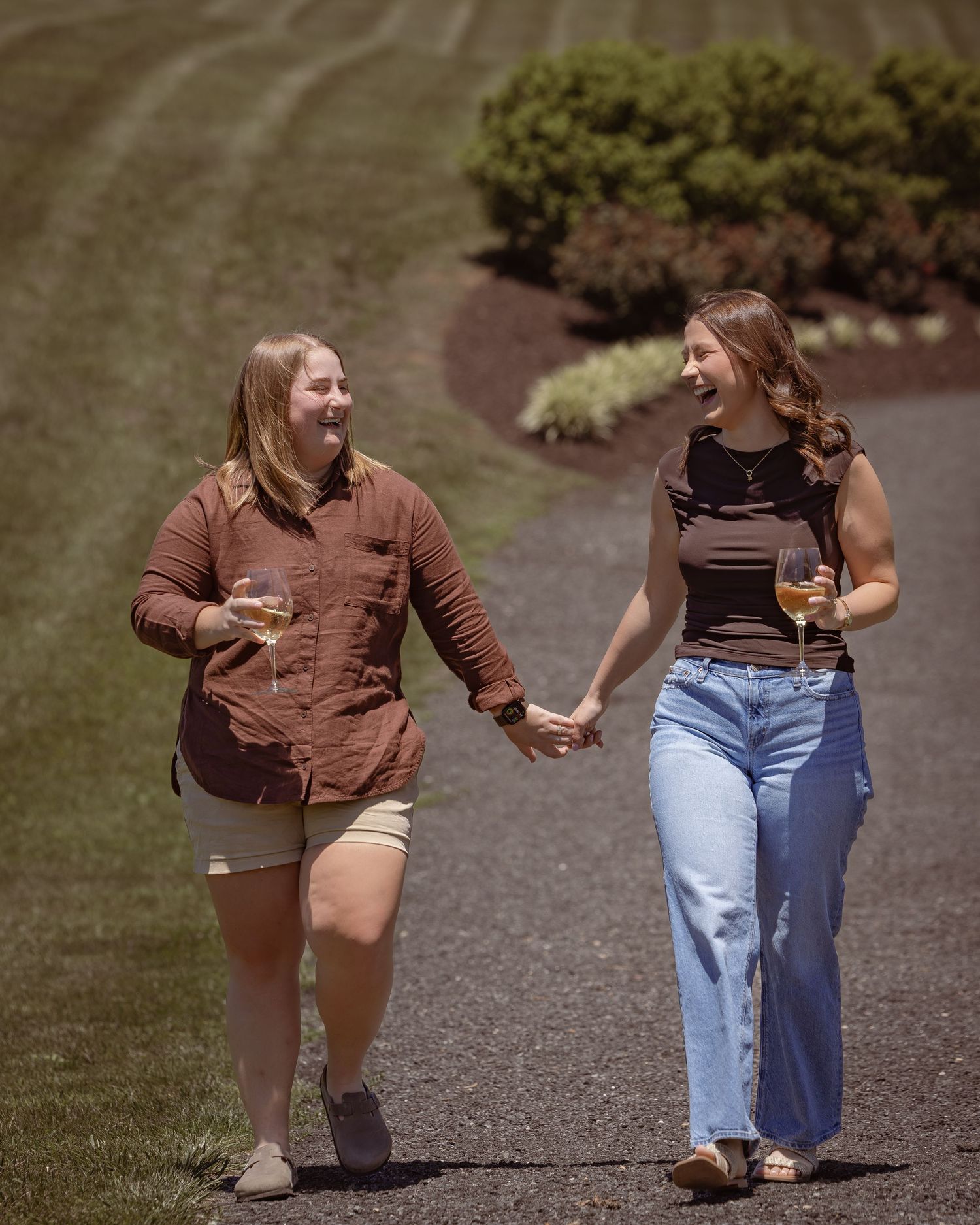 Two women holding hands and laughing while walking outdoors on a sunny day, each holding a glass of wine.