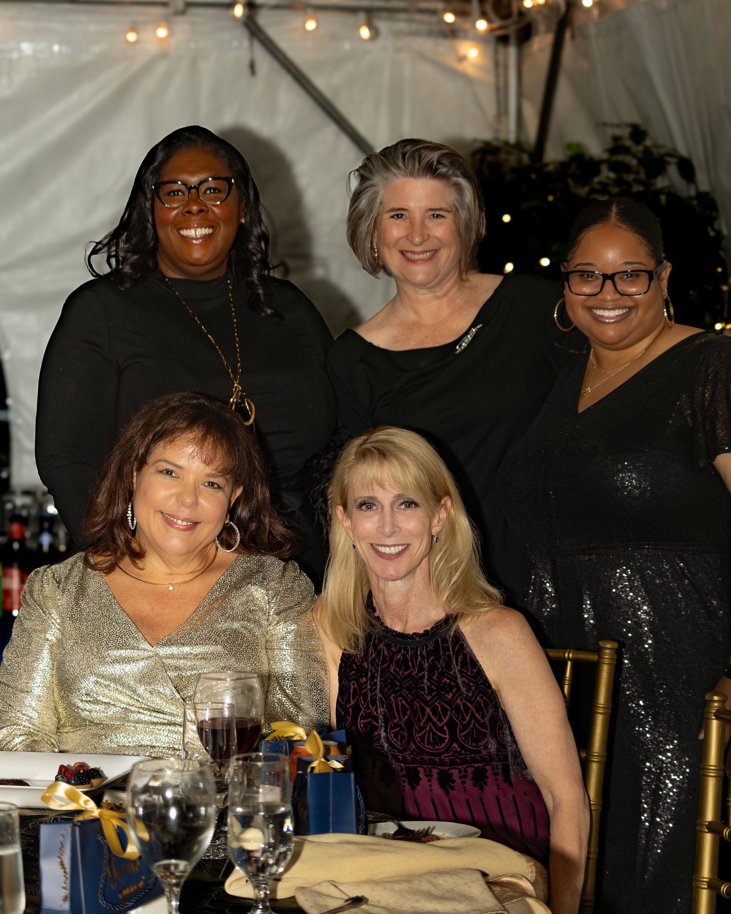 A group of friends enjoying dinner and drinks together at a festive evening gathering with string lights overhead.