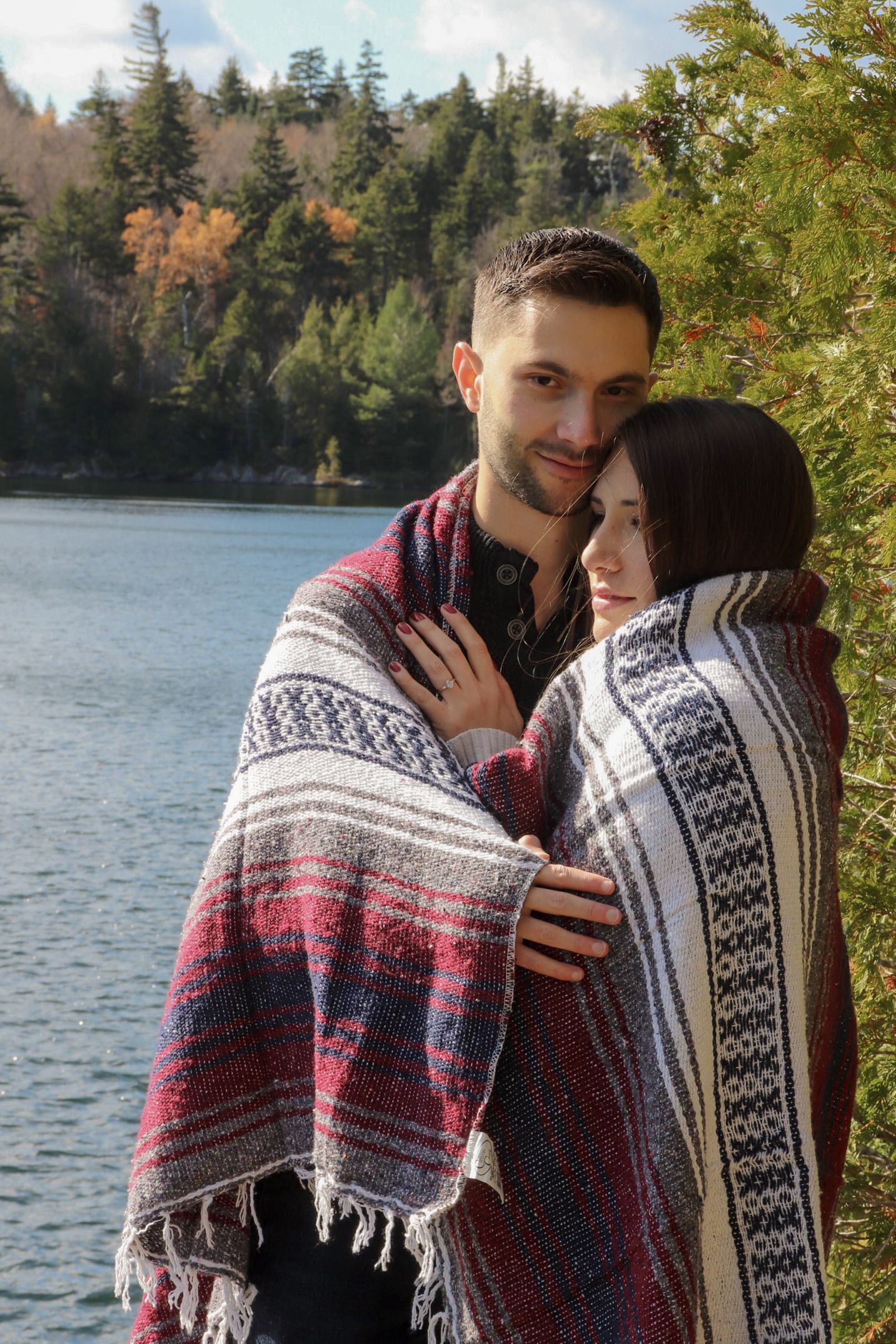 Happy couple wrapped in a cozy plaid blanket embracing outdoors near a lake surrounded by autumn trees.