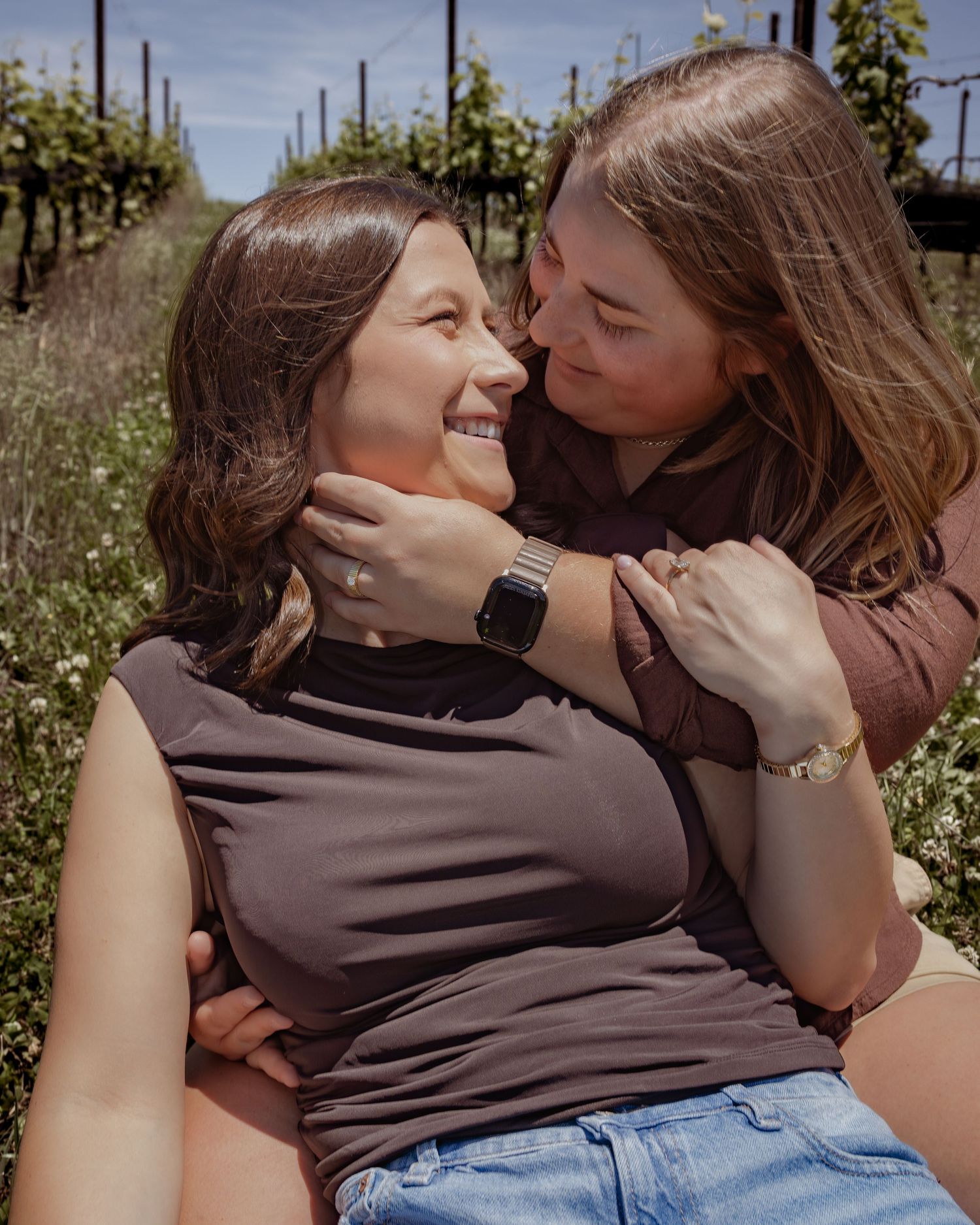 Two women share a joyful moment sitting in a sunny vineyard, one smiling up as the other embraces her from behind.