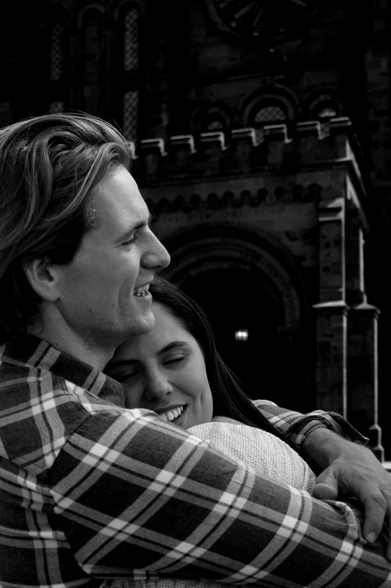 Black and white photo of engaged couple embracing and laughing in front of Smithsonian Castle.