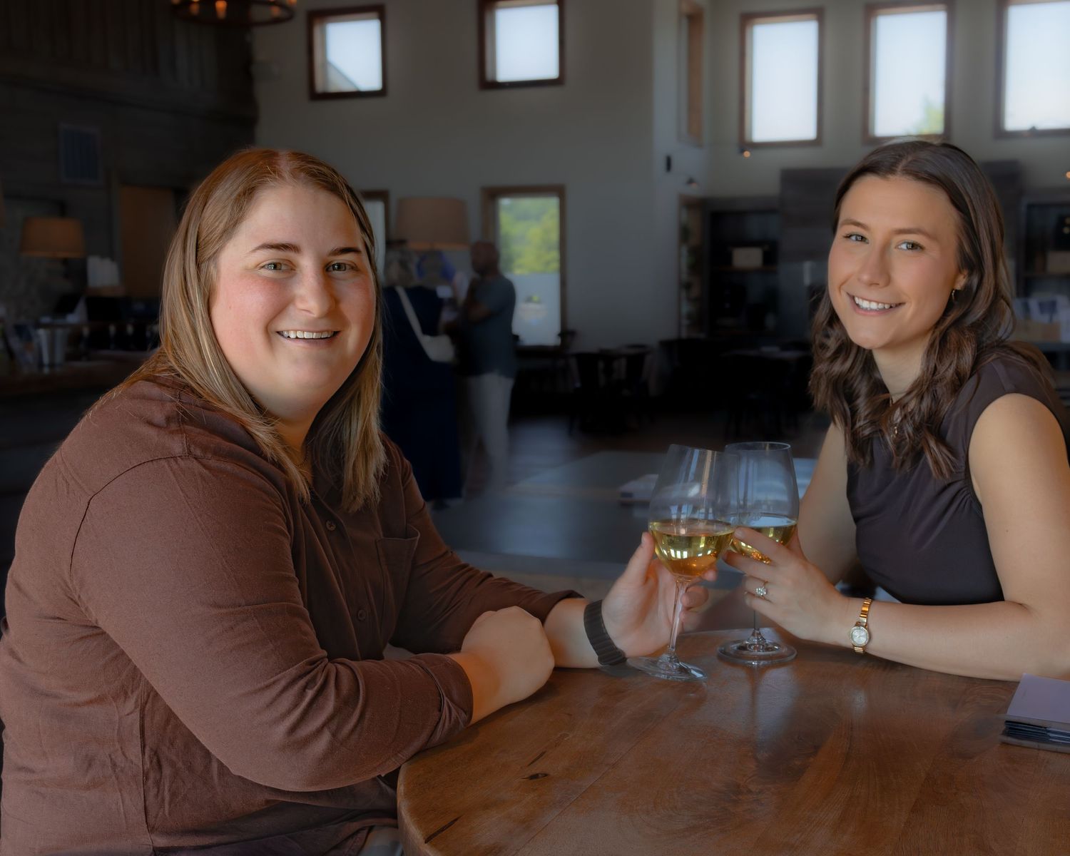 Two women smiling and toasting with wine glasses at a wooden bar inside a modern winery tasting room.