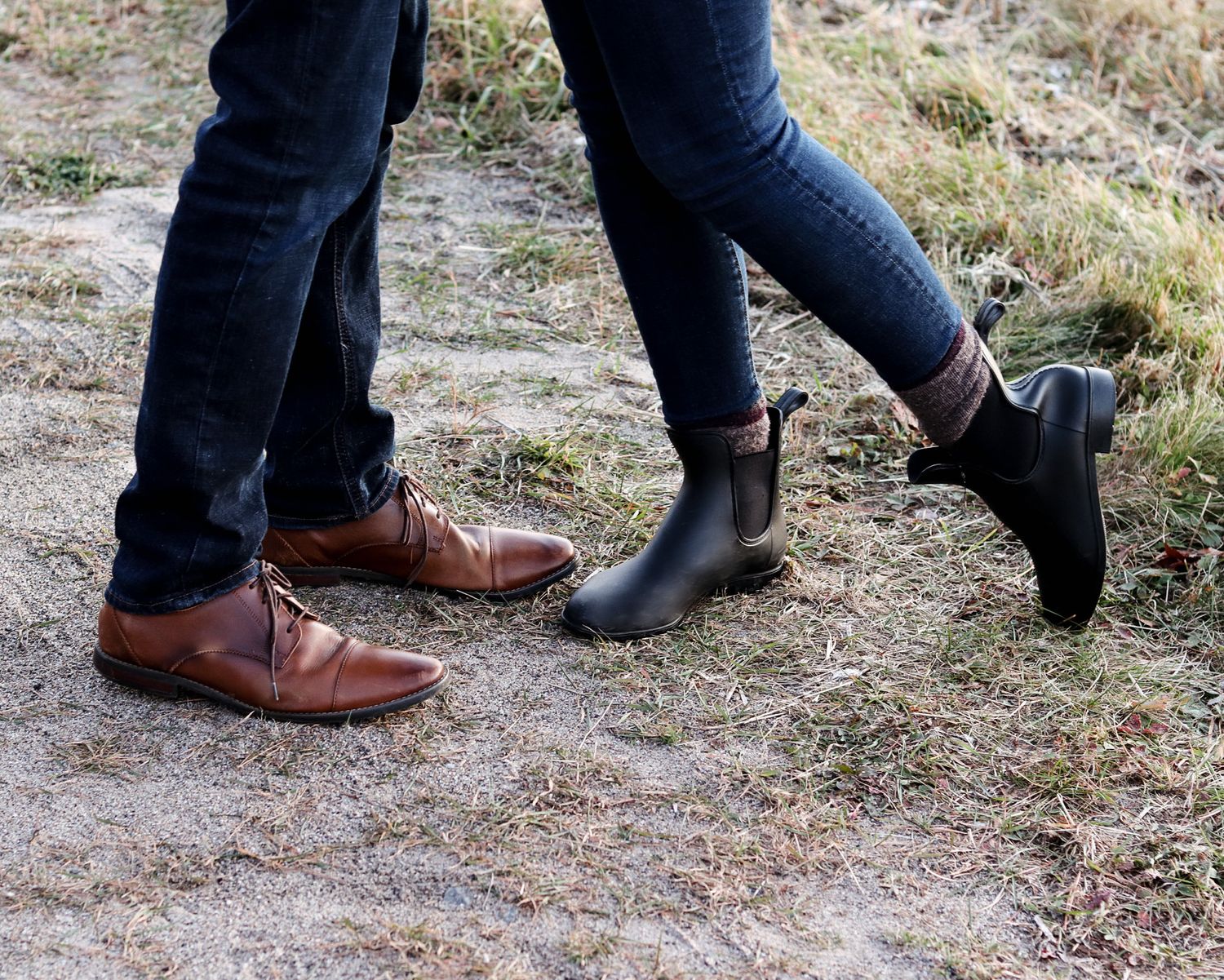 Two people standing close together outdoors, one wearing brown leather lace-up shoes and the other in black Chelsea boots on grassy ground.