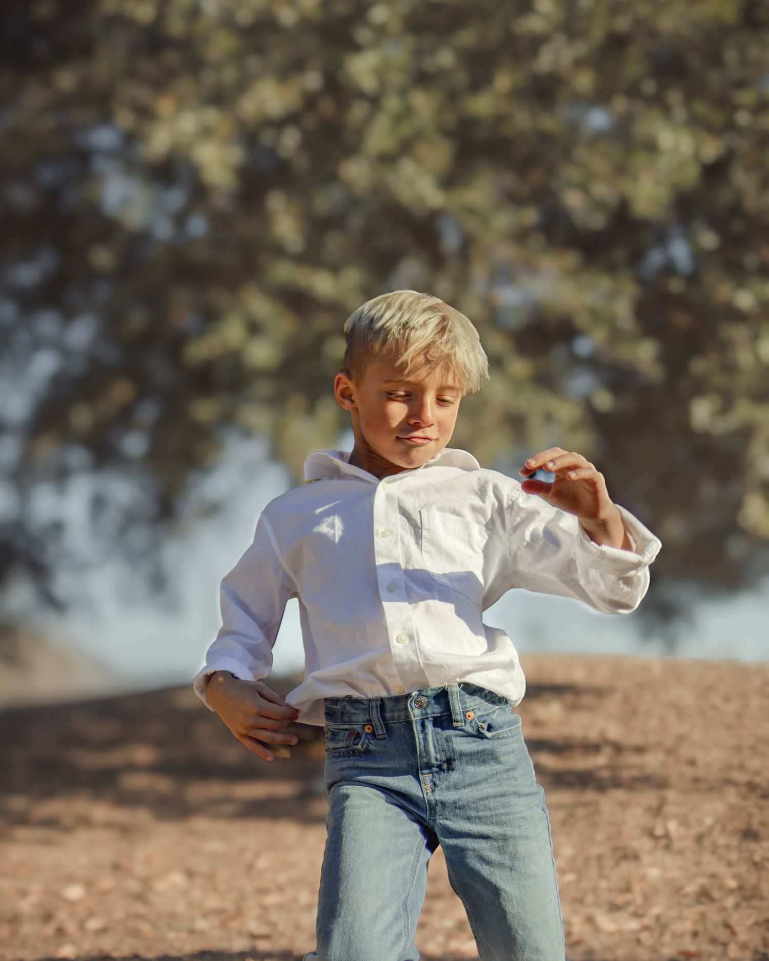 Young child in a white button-down shirt and jeans dances playfully outdoors on a sunny day with trees in the background.
