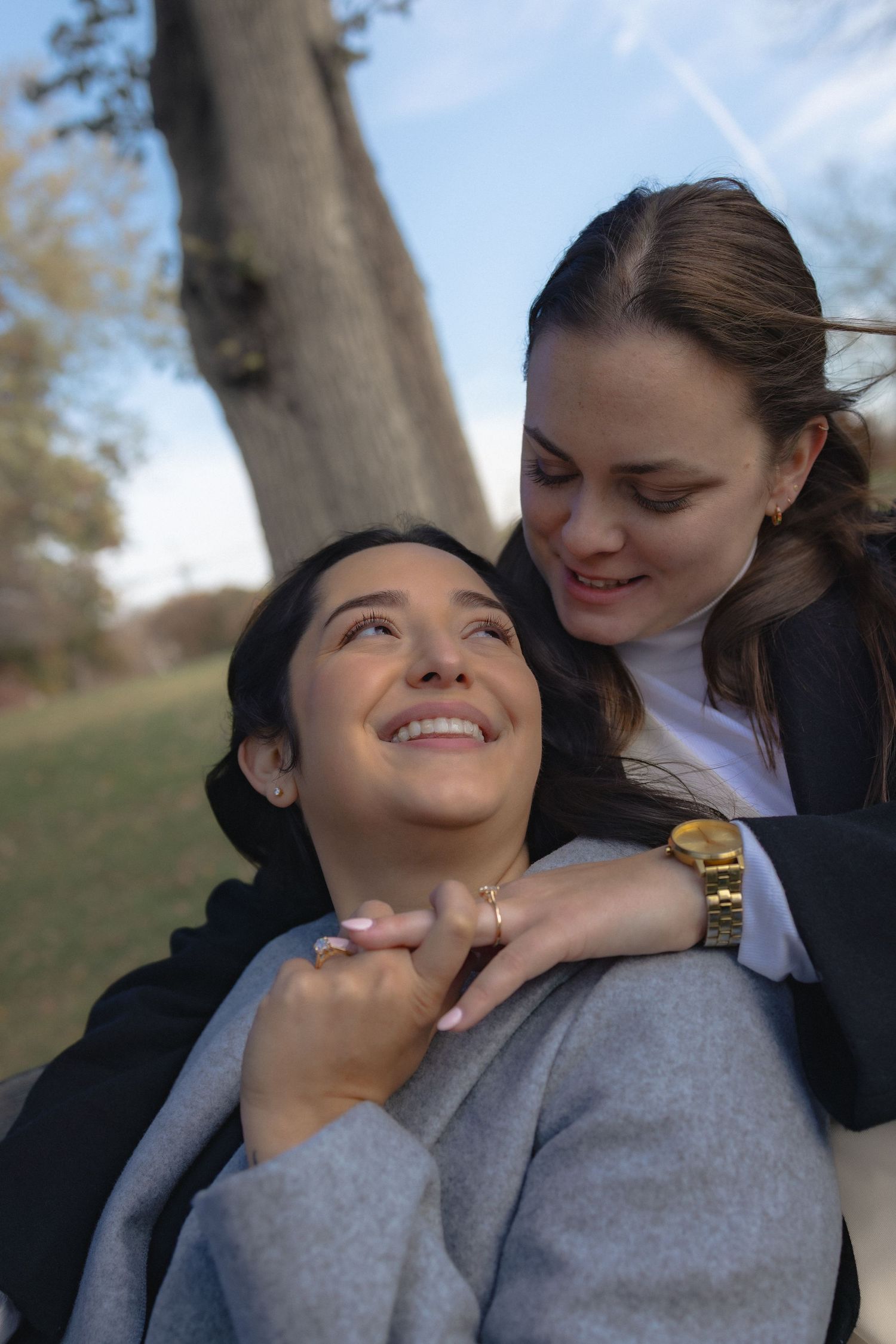 Two friends share a tender moment outdoors, smiling and embracing while relaxing against a tree on an autumn day.
