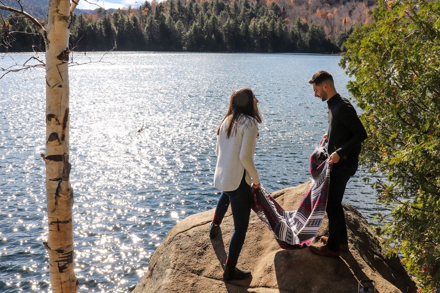 Couple standing on a rocky lakeside cliff surrounded by autumn trees and sparkling sunlit water.