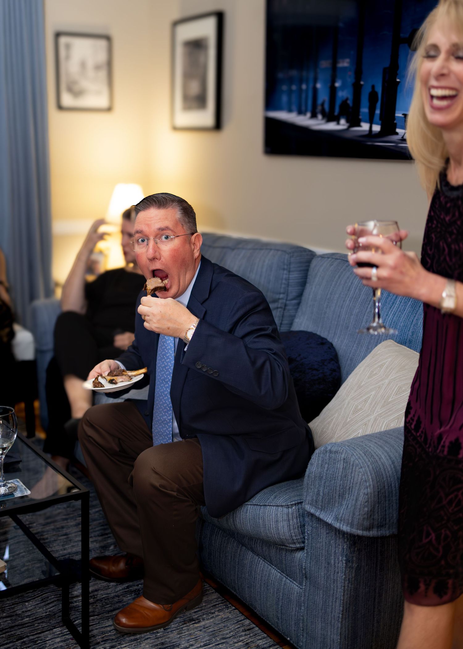 A person in a suit sits on a gray couch while laughing and gesturing during what appears to be a social gathering.