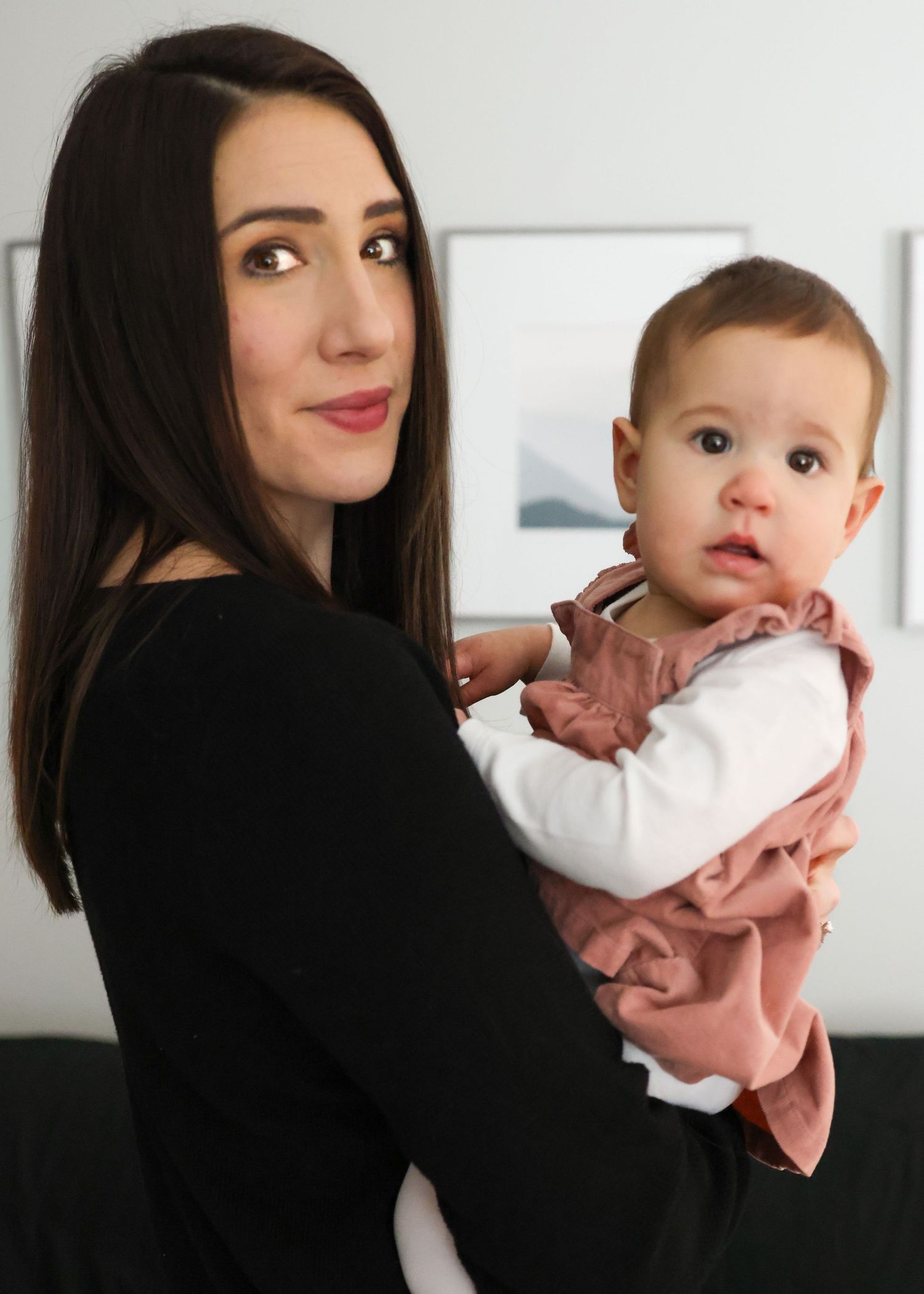 A parent in a black top holds a baby wearing a white and pink outfit against a light-colored wall.