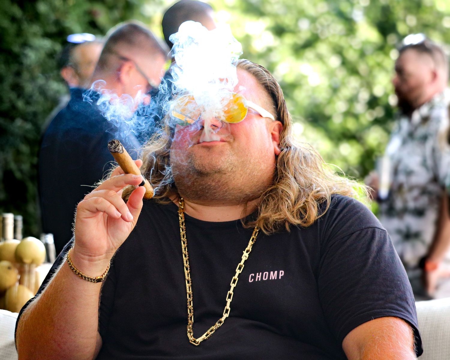 A person wearing a black t-shirt blows smoke from their mouth while wearing a gold chain necklace outdoors.