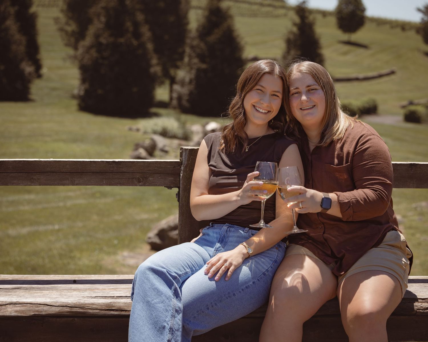 Two women smiling and toasting with wine glasses on a sunny bench with green hills in the background.