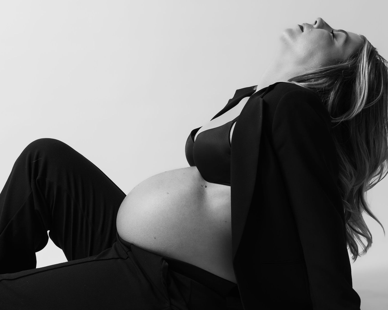 Black and white photo of a pregnant woman in a black blazer and bra, reclining gracefully with her bare bump exposed.