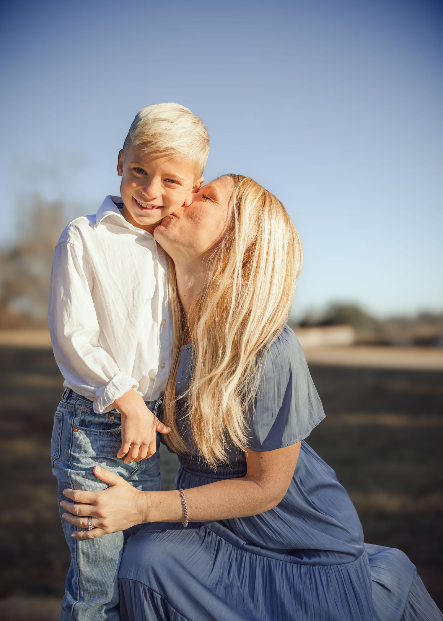A blonde mother in a blue dress kisses her smiling blonde son outdoors in a sunny field.