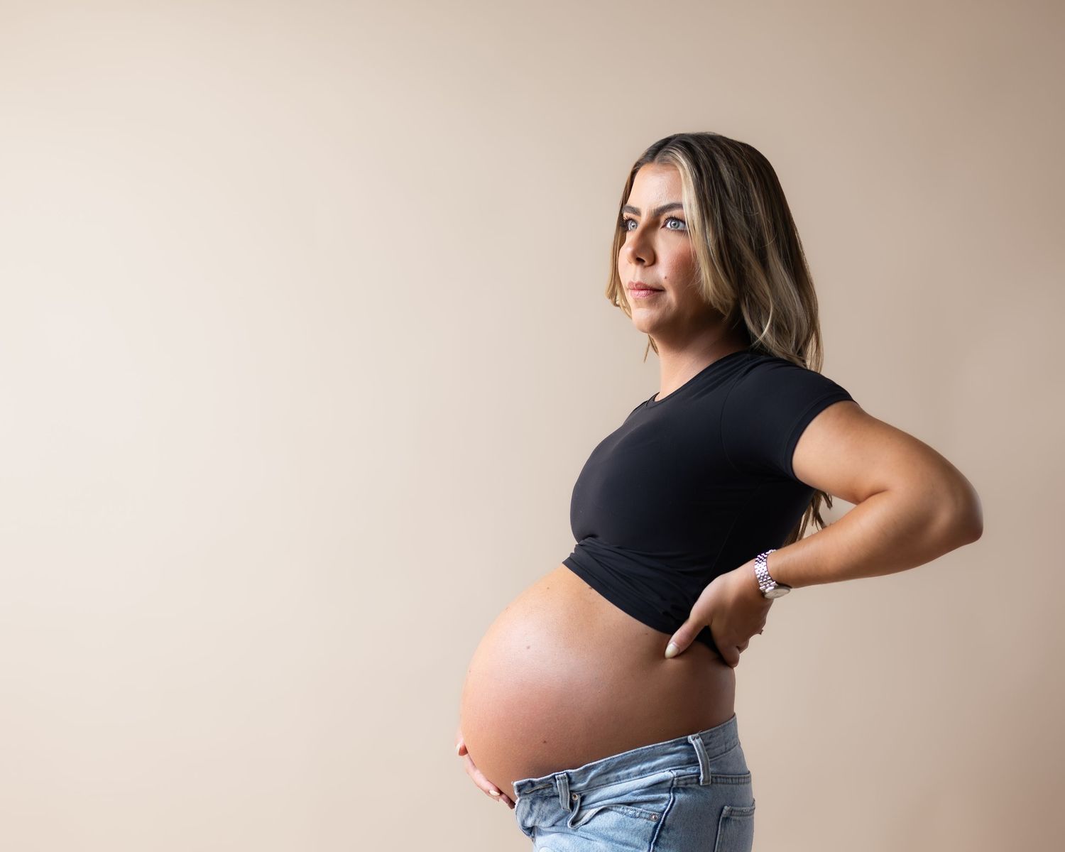 Pregnant woman in black crop top and jeans posing sideways showing baby bump against beige background.