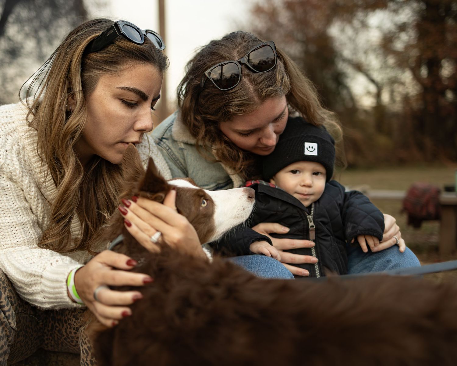 Two women and a baby interact with a brown and white dog outdoors on a fall day.