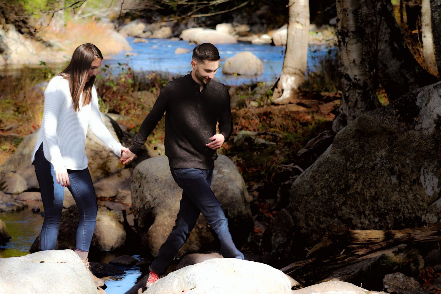 Couple holding hands while carefully stepping over rocks near a forest stream on a sunny day.