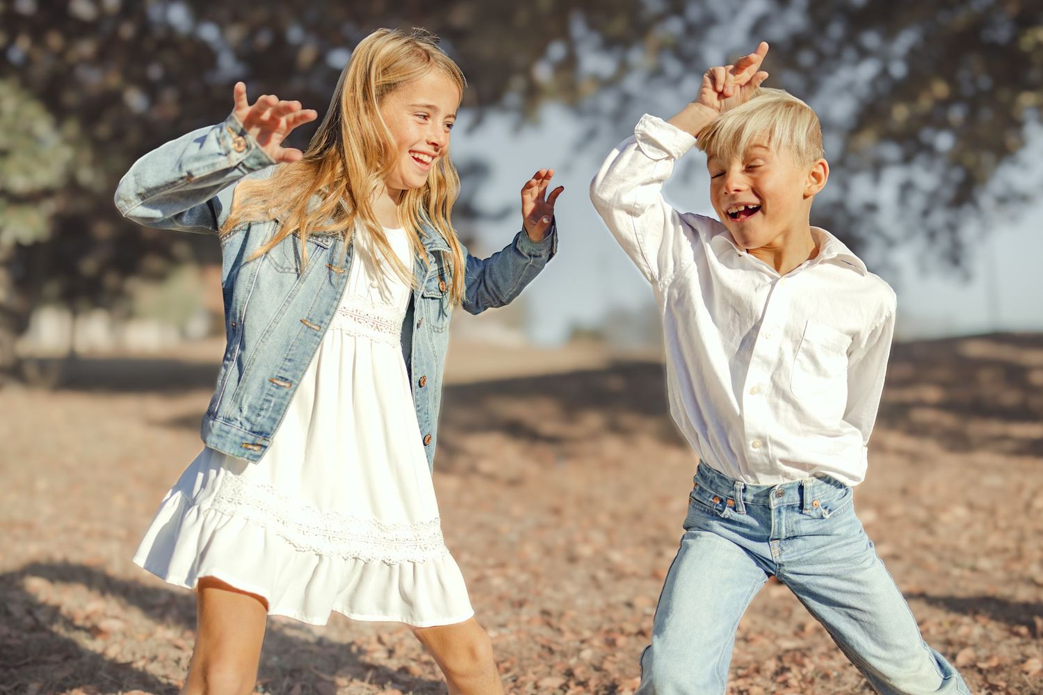 Two happy children dancing outdoors in casual denim outfits, laughing and playing together in a sunny park setting.