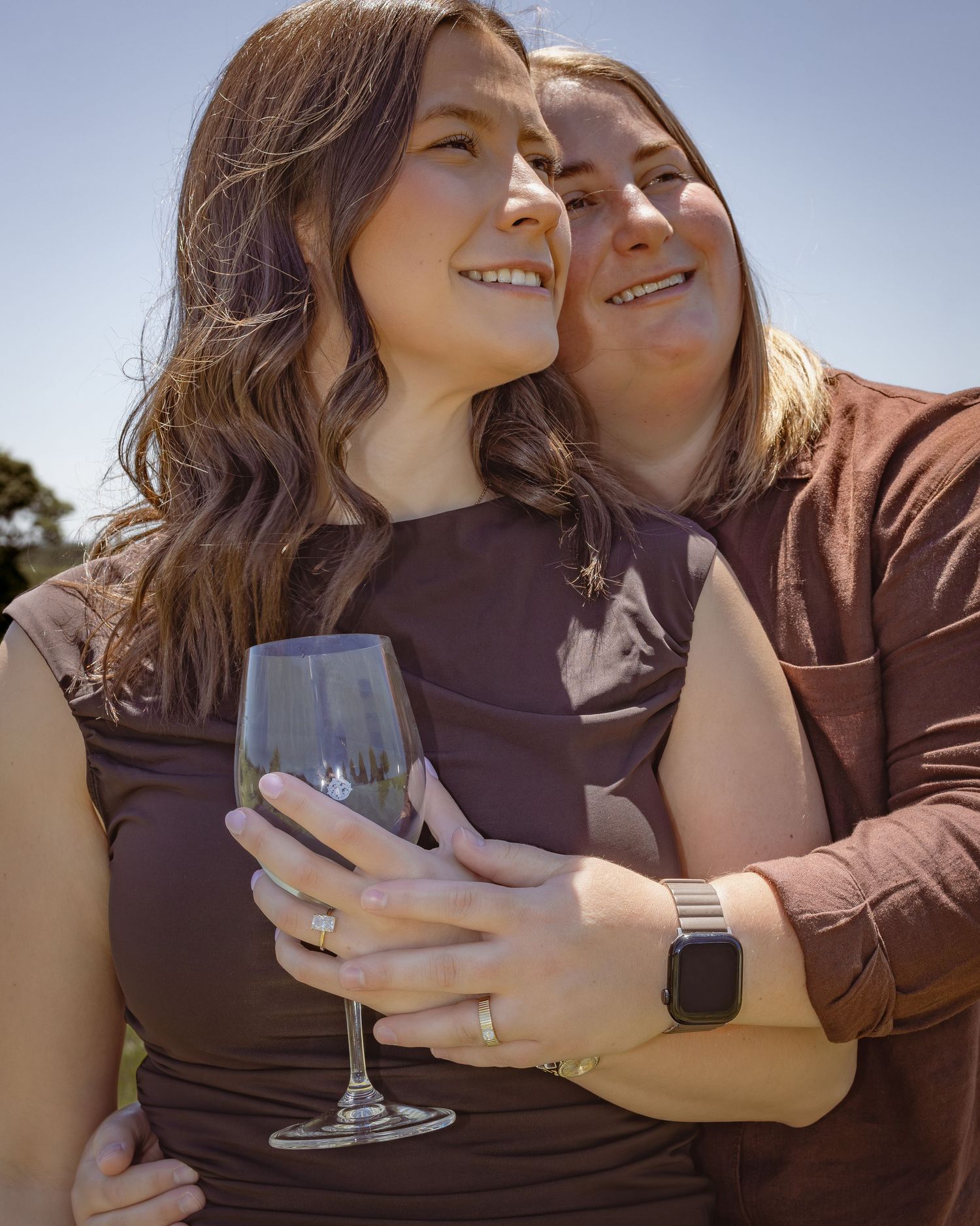 Two women in matching brown outfits embrace outdoors, one holding a wine glass, smiling joyfully on a sunny day.