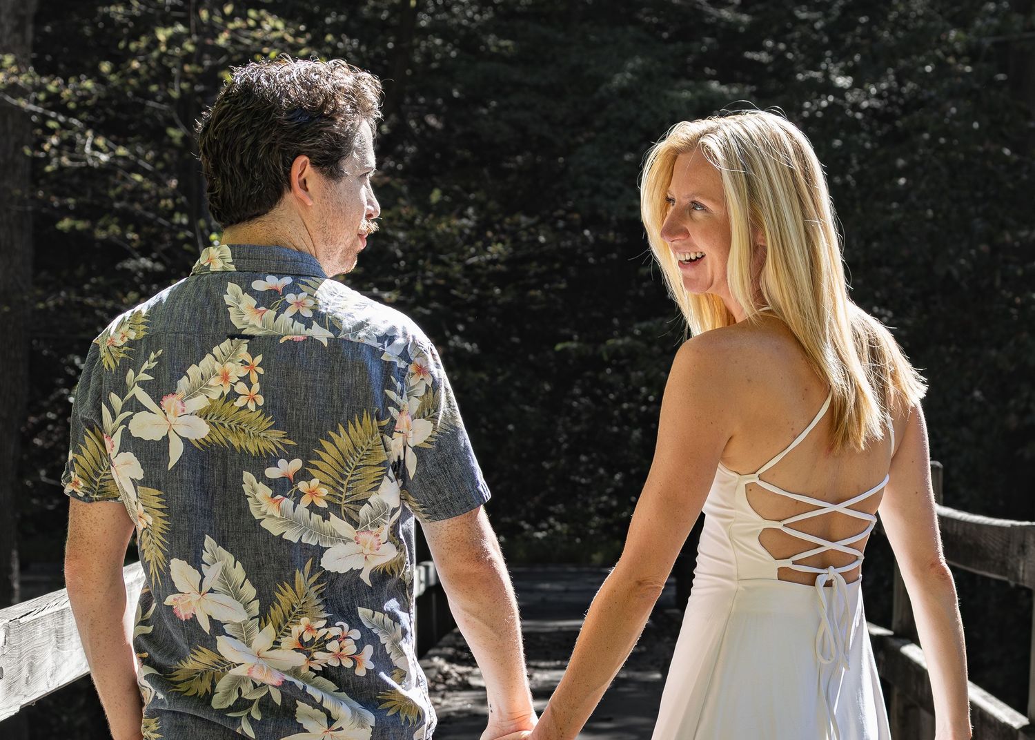 A couple holding hands while walking outdoors, with one wearing a floral shirt and the other in a white strappy dress.