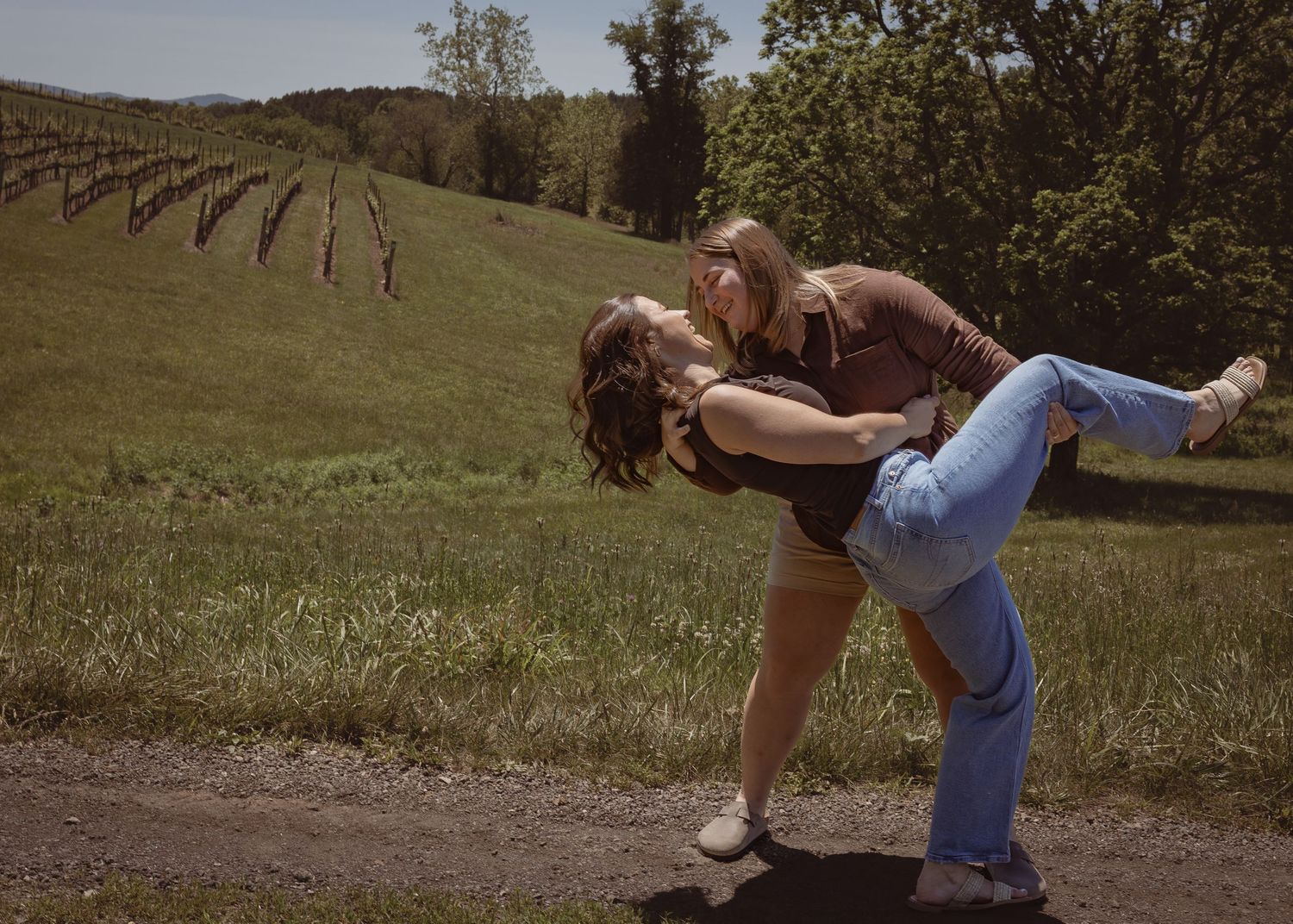 Couple sharing a romantic dip pose on a dirt path beside a lush green vineyard on a sunny day.