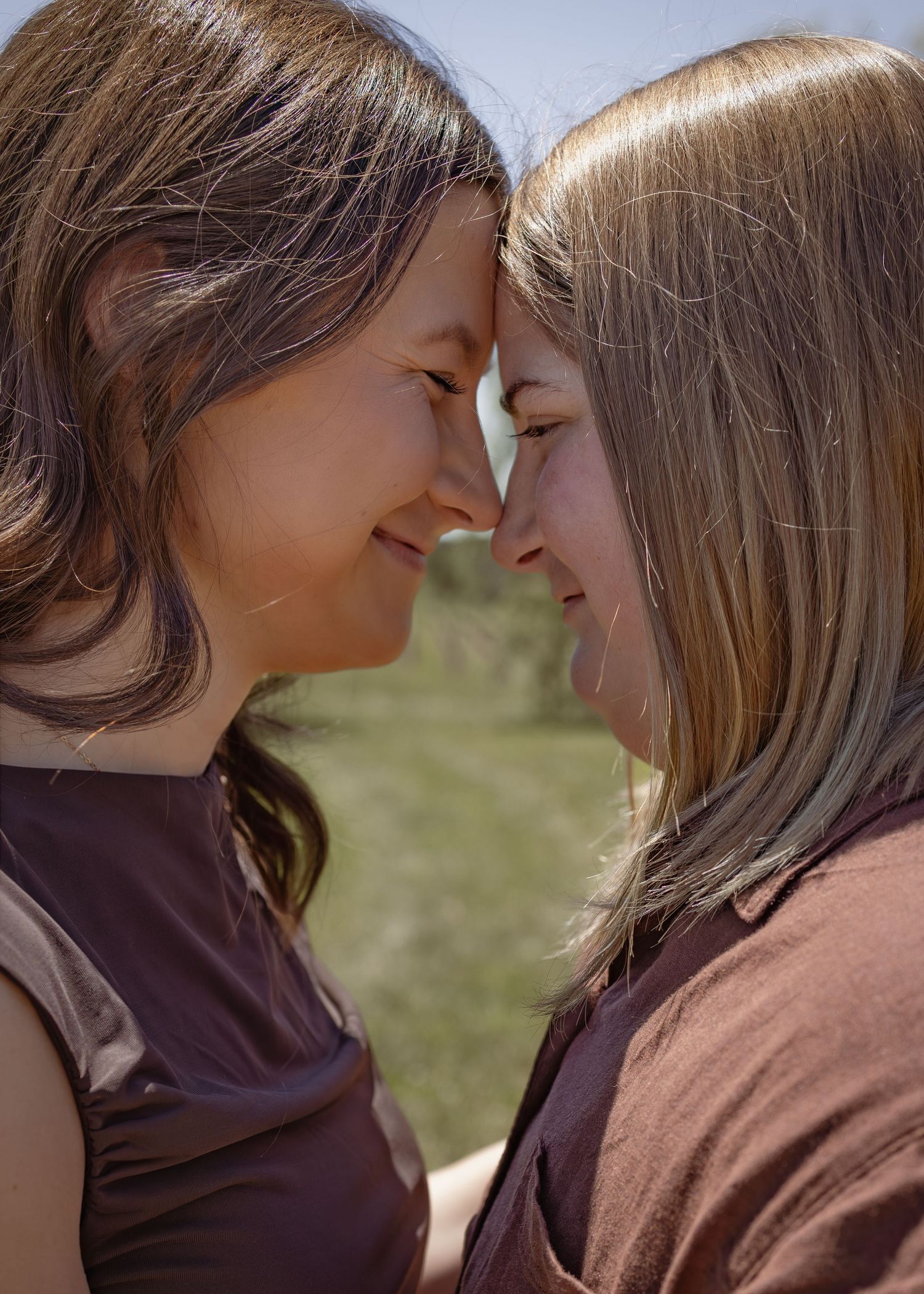 Two fiancees touching foreheads and smiling warmly at each other outdoors on a sunny day.