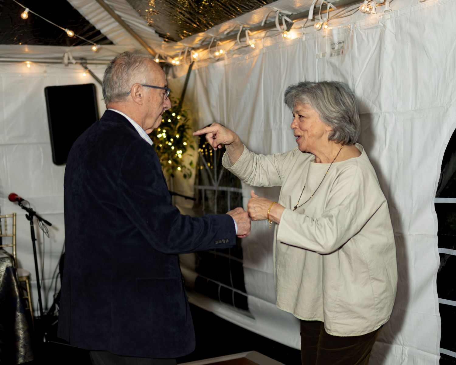 Two people interact animatedly under string lights in what appears to be a tent or covered outdoor space.