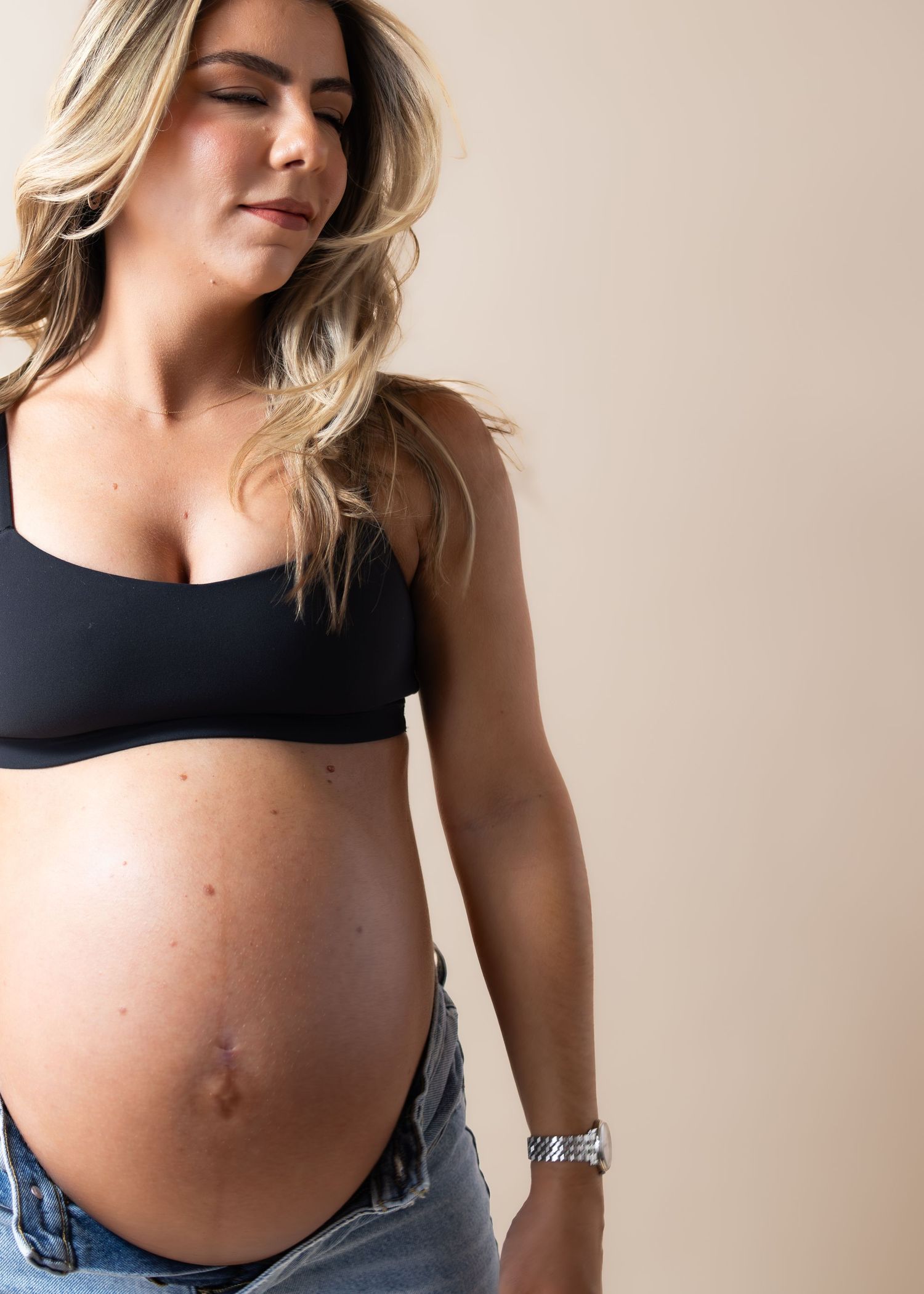 Pregnant woman in black crop top and denim jeans showing baby bump, looking down with blonde hair.