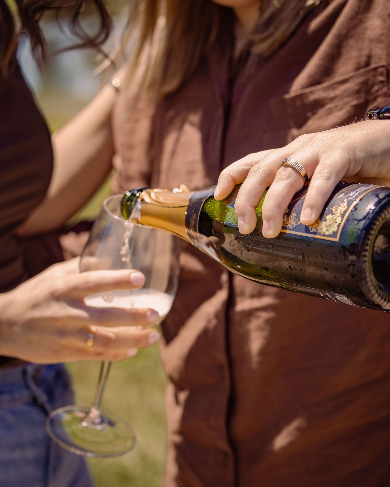 Person pouring sparkling wine from a bottle into a glass outdoors on a sunny day.