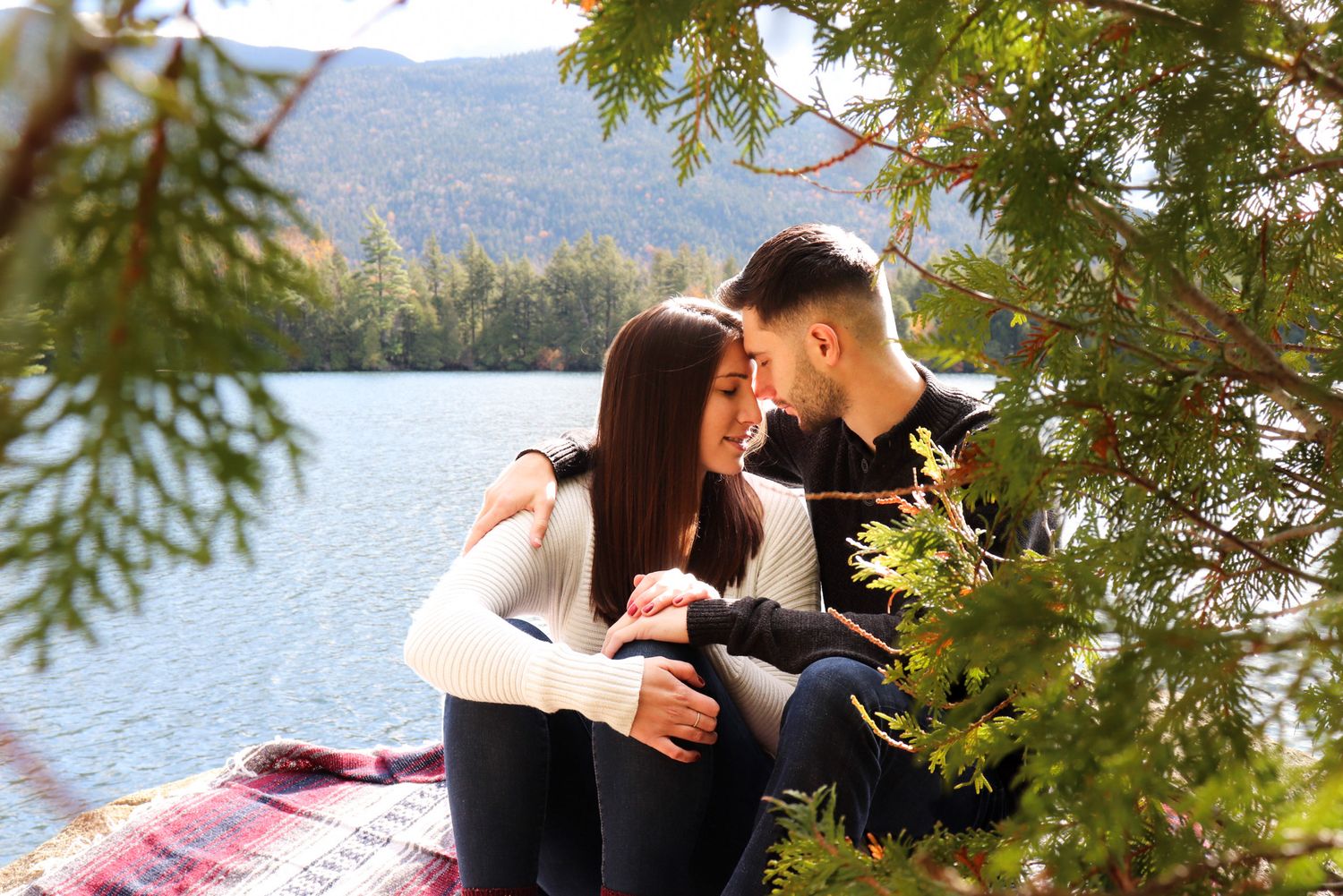 A young couple sits intimately by a mountain lake, framed by evergreen branches on a plaid blanket.