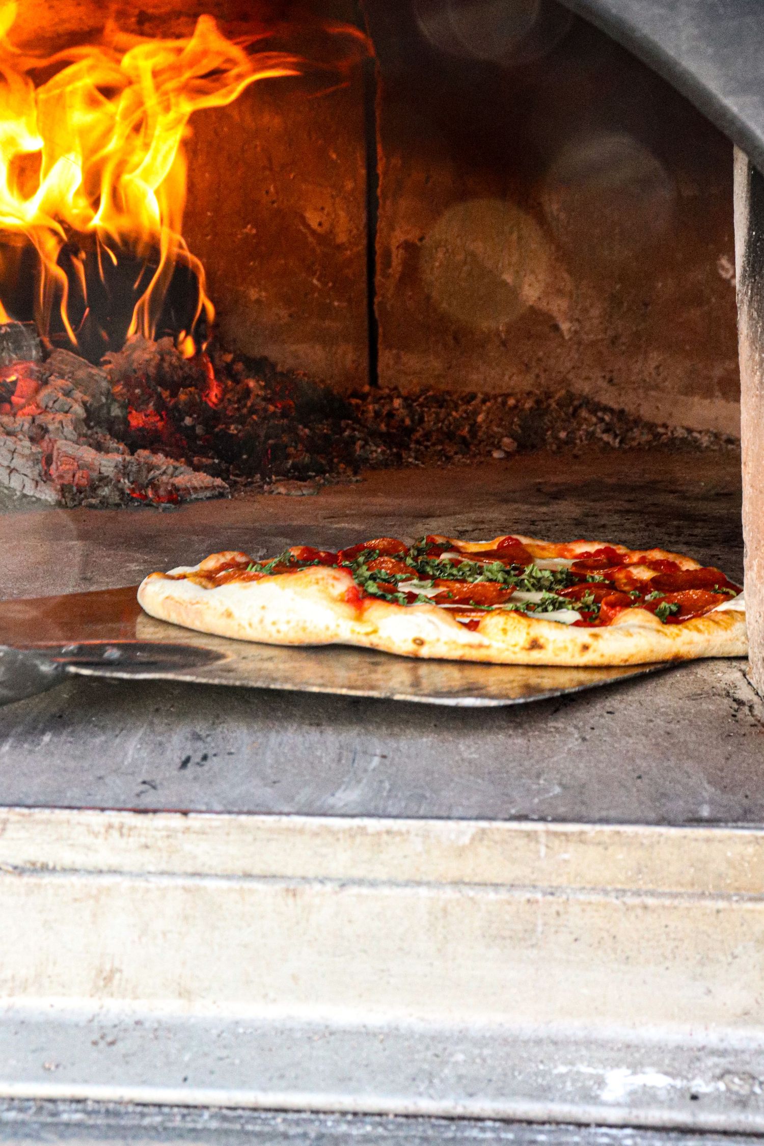 Fresh pizza with toppings cooking in a traditional wood-fired brick oven with roaring flames in the background.