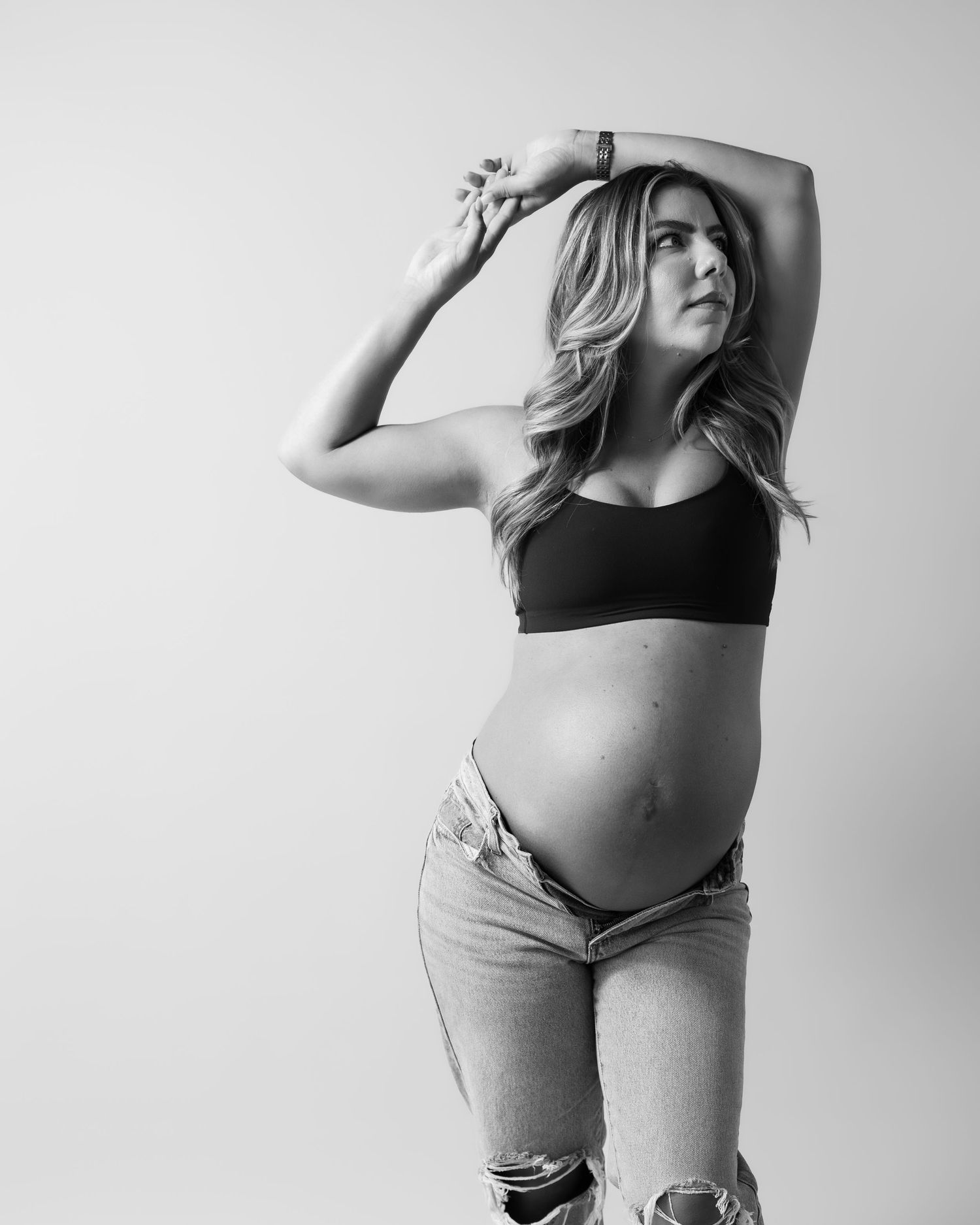 Black and white maternity photo of a pregnant woman in a crop top and jeans posing with arms raised.