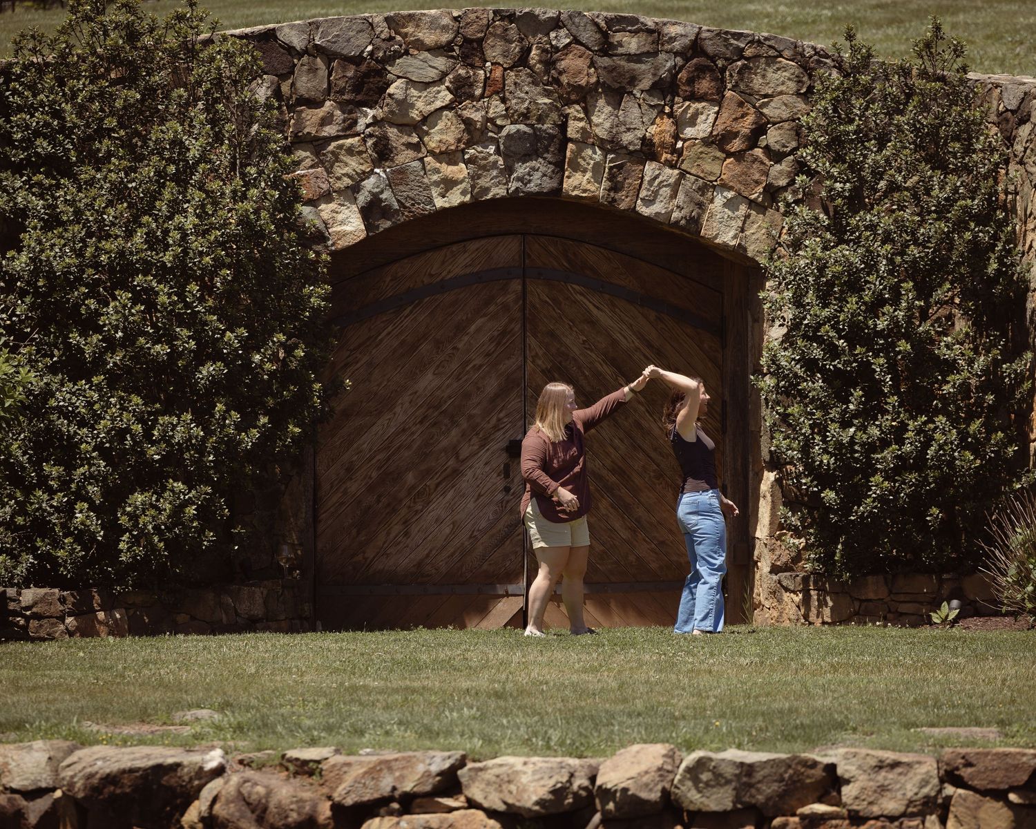 Couple dancing outdoors in front of a rustic stone arch and wooden door surrounded by lush green hedges.