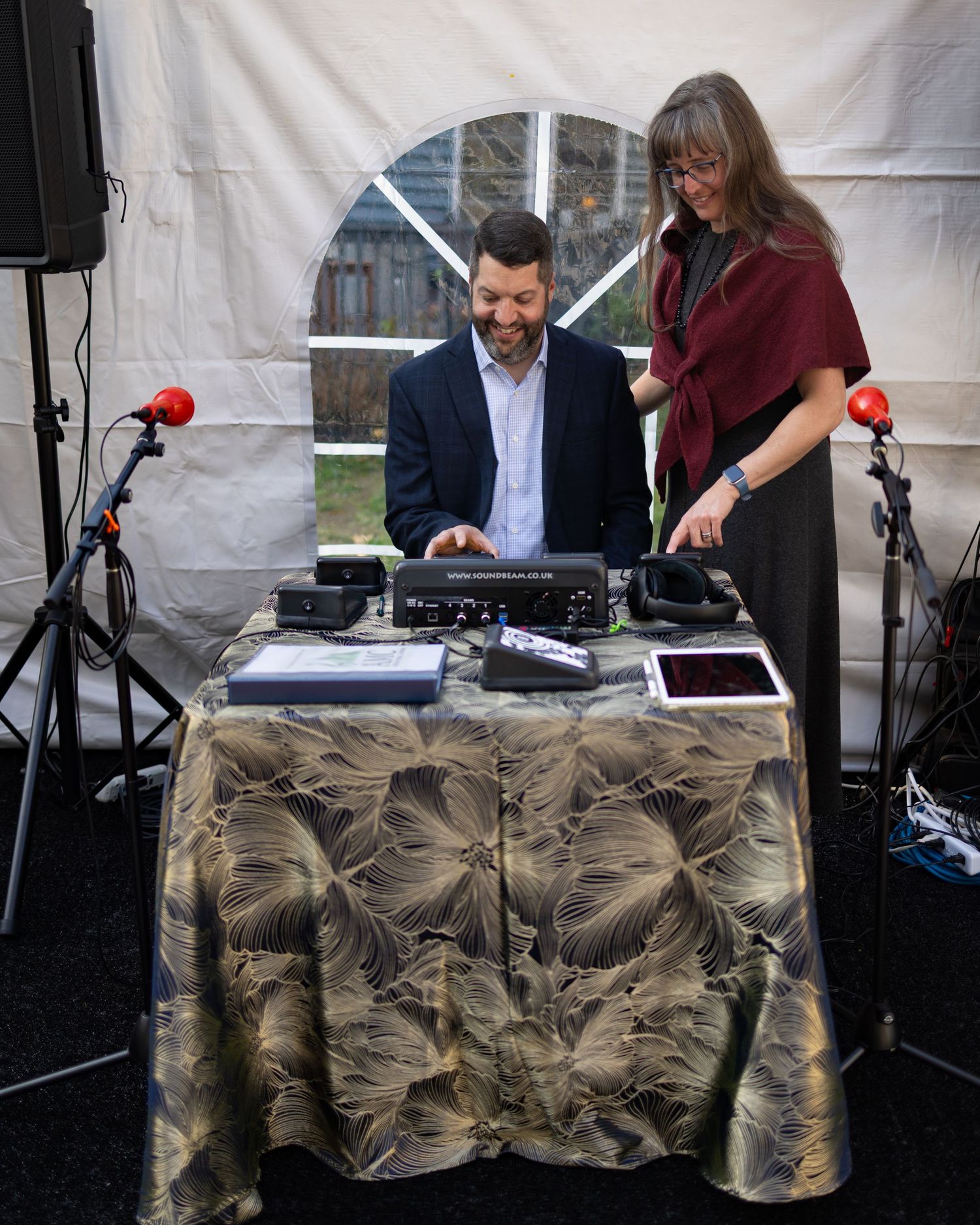 Two people operate DJ equipment and audio gear at an event setup with microphones and a decorative tablecloth.