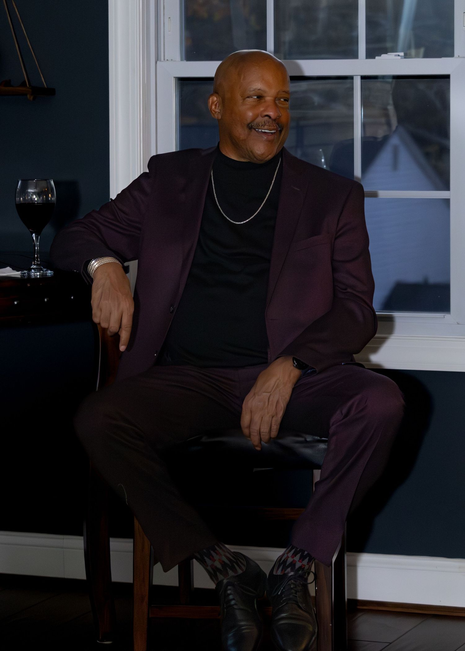 Person in burgundy suit and black turtleneck sits relaxed in wooden chair near window with wine glass in background.