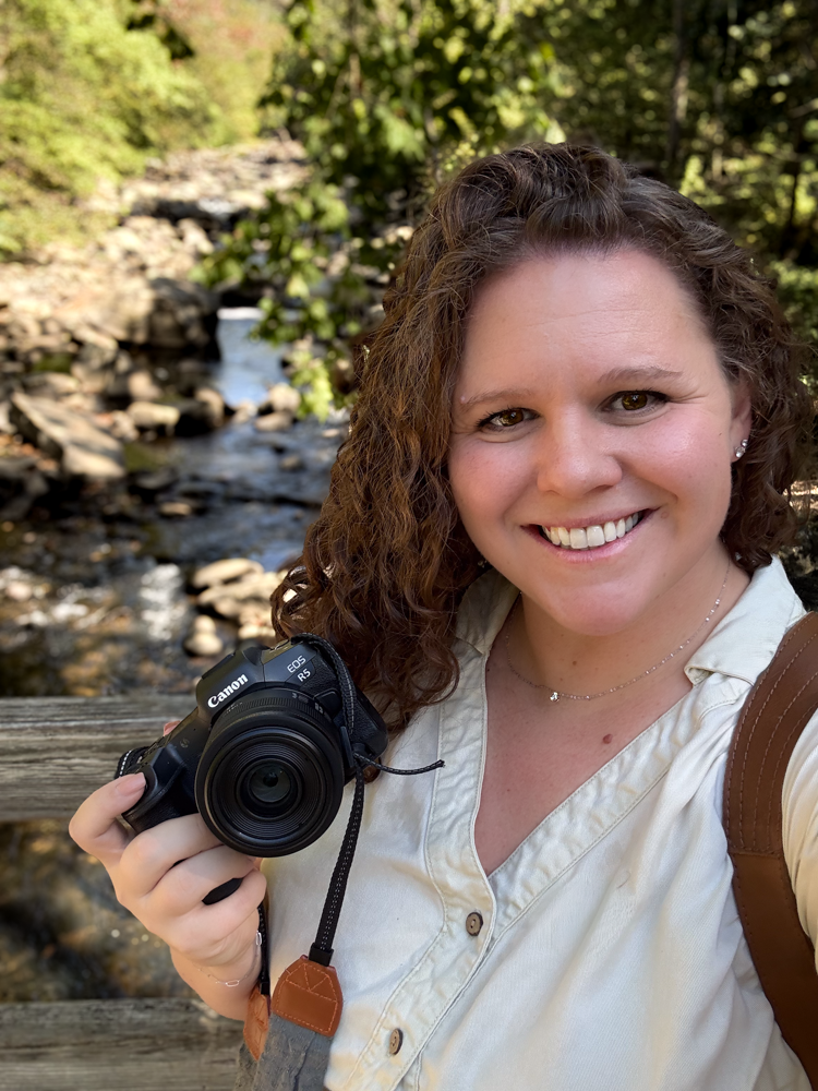 A photographer with curly hair and a white shirt holds a camera while smiling outdoors by a stream in natural lighting.