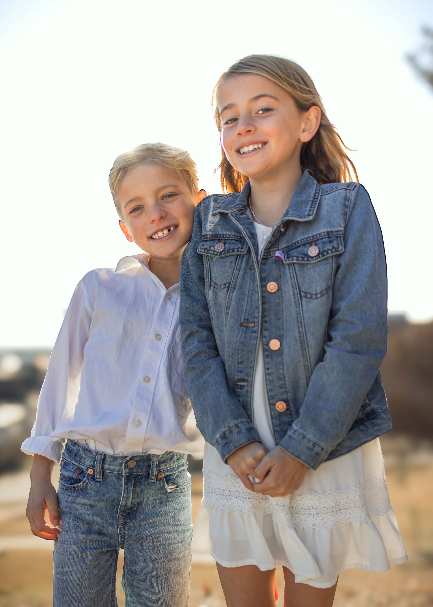 Two smiling children pose outdoors, girl in denim jacket and white dress, boy in white shirt and jeans.