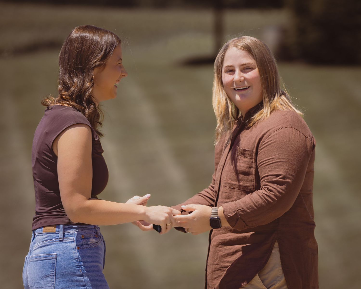 Two smiling women holding hands outdoors, wearing brown tops and jeans, sharing a joyful moment together.