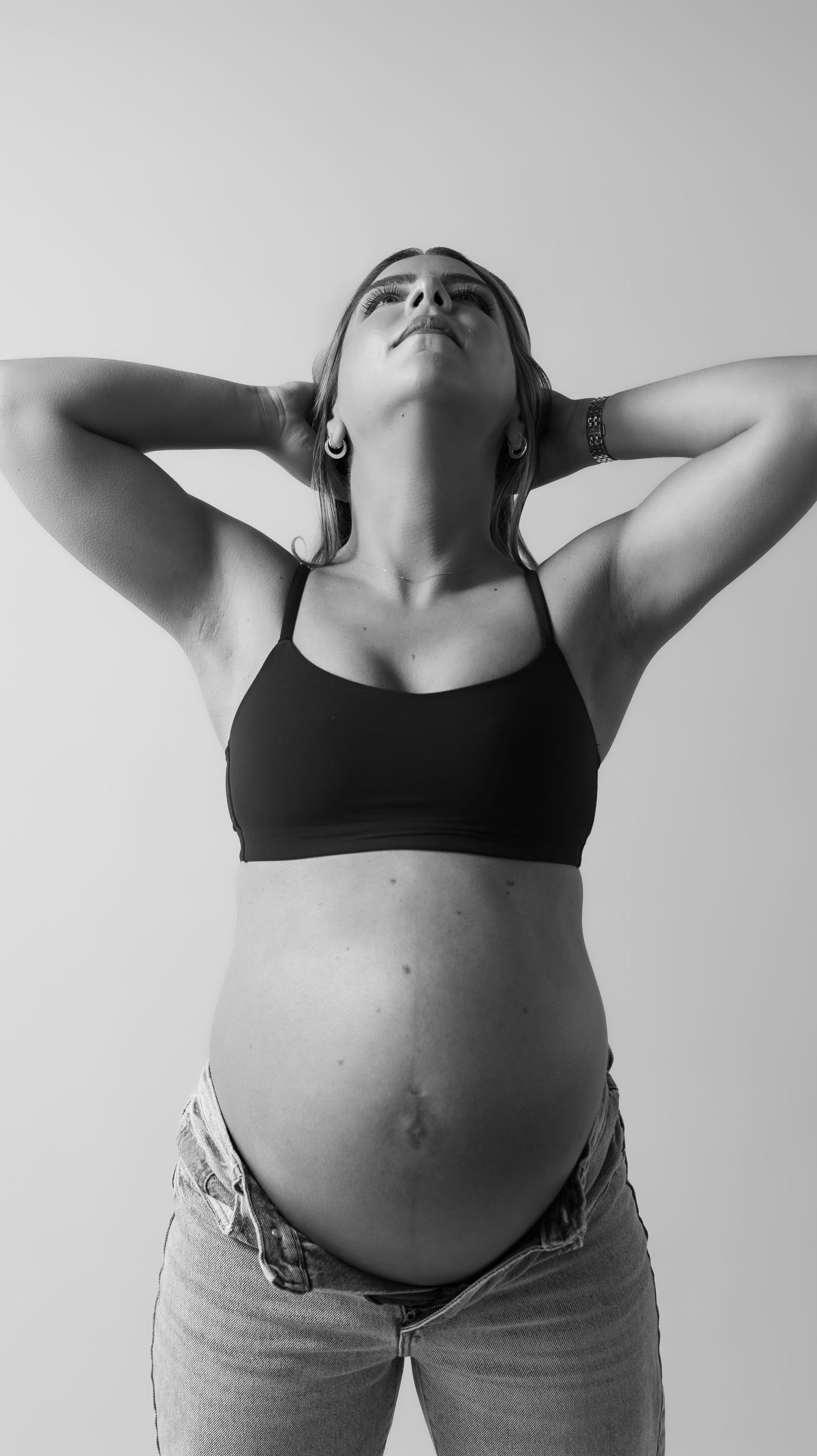Black and white photo of a pregnant woman in a crop top and jeans, hands behind her head, looking up.