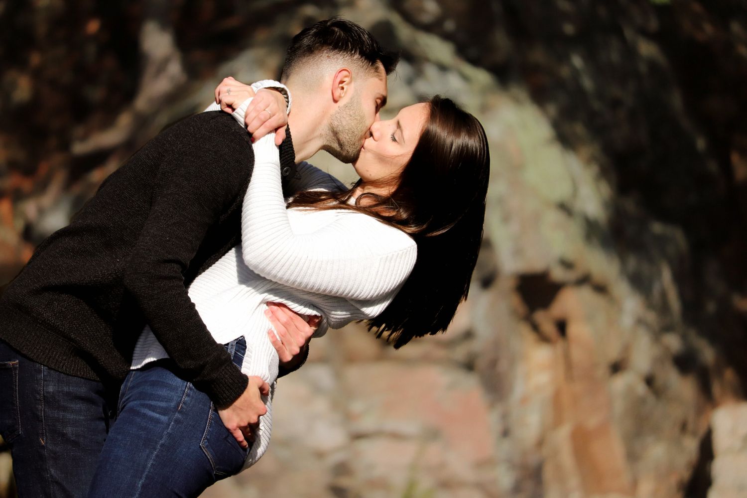 Romantic couple sharing a tender kiss outdoors near rocky background in casual attire.