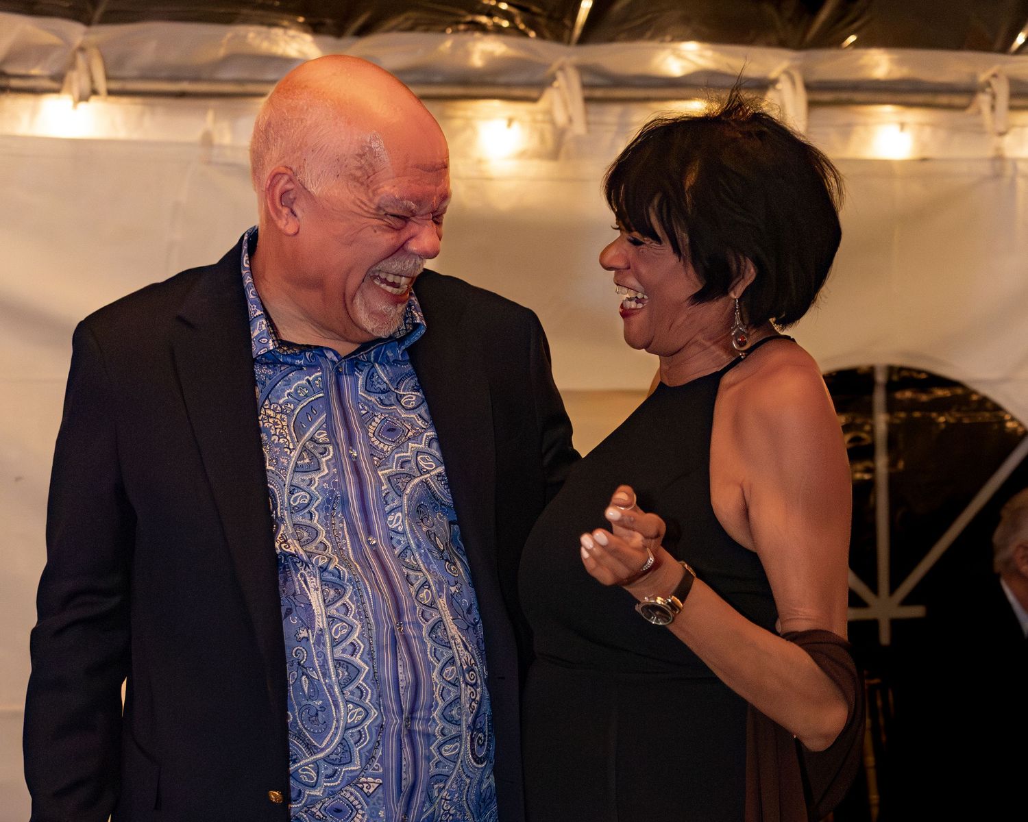 A person in a blue shirt and dark jacket interacts with others at an evening event with string lights overhead.