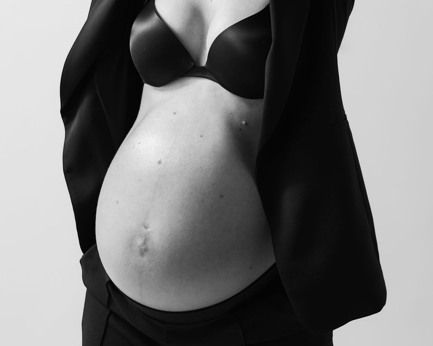 Black and white maternity photo showcasing a pregnant woman's bare baby bump, wearing a black bra and blazer.