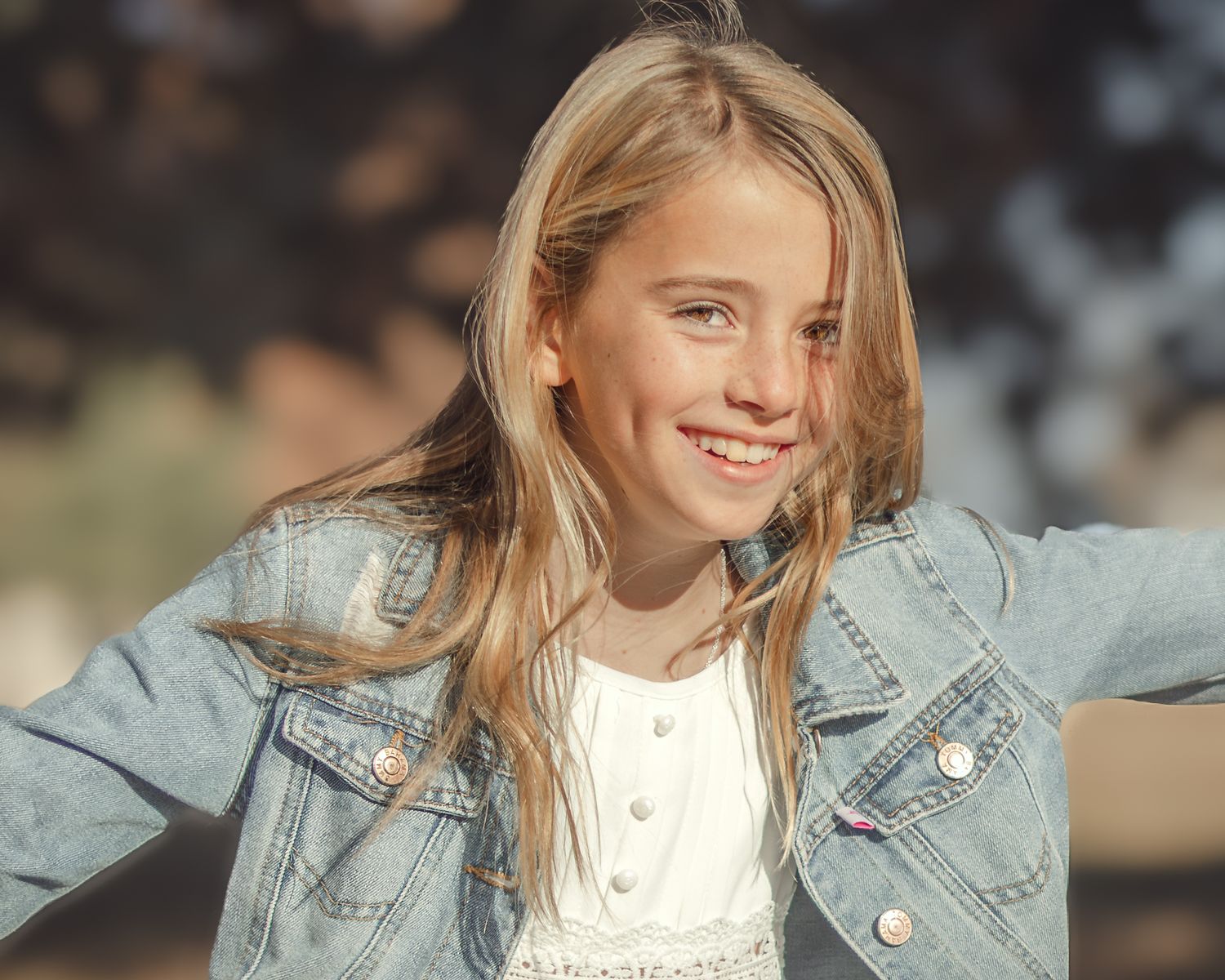 A person in a light denim jacket and white top smiles while posing against a blurred background.