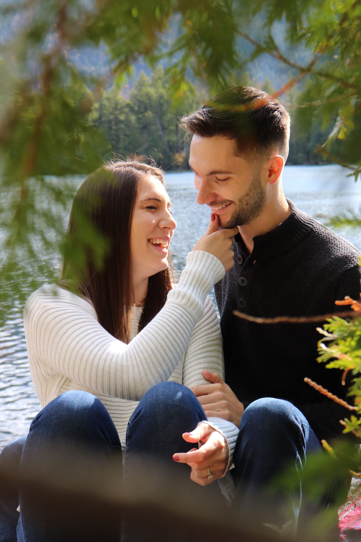 Happy couple sitting by a lake, smiling at each other, woman touching mans face, wearing casual sweaters outdoors.