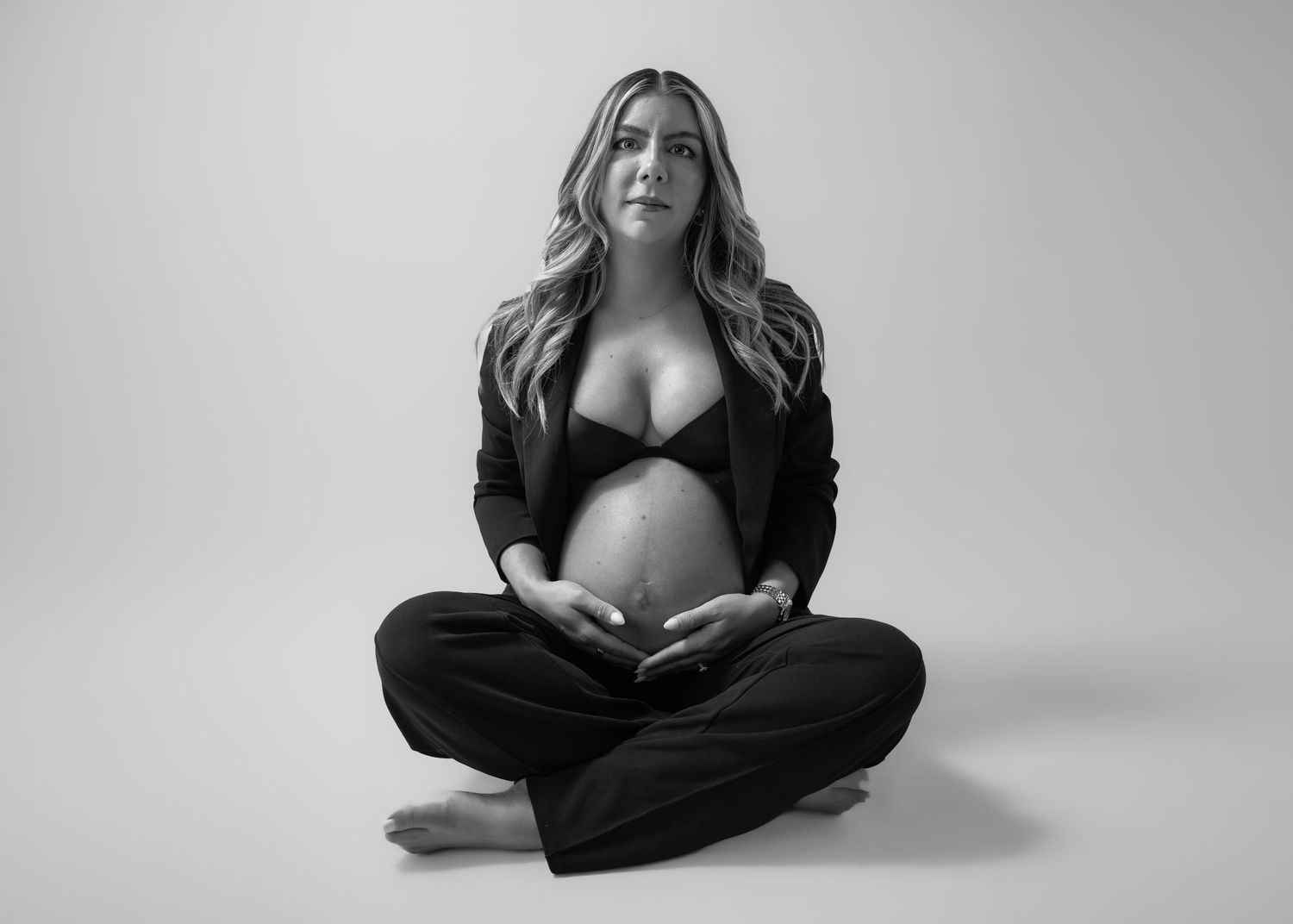 Black and white maternity photo of a woman in a blazer and bra sitting cross-legged on the floor cradling her baby bump.