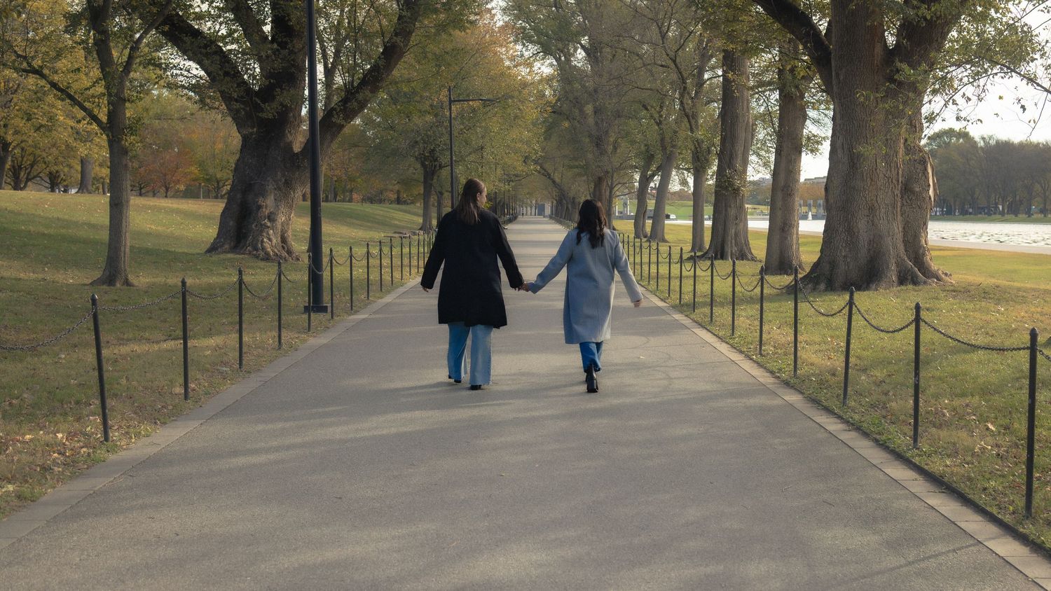 Two women walk hand in hand along a tree-lined park path on the National Mall on an autumn day.