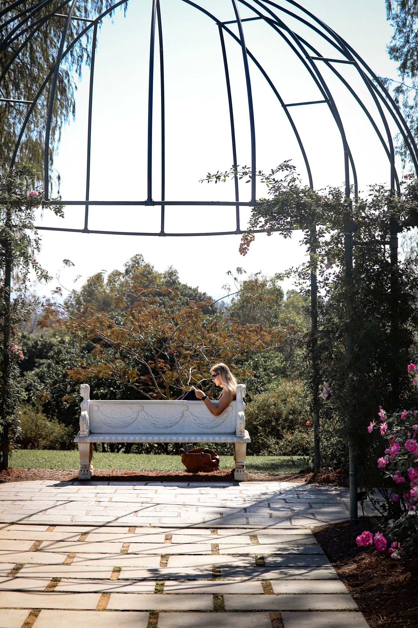 A person lounges on a white bench in a serene garden gazebo surrounded by lush trees and flowering plants.