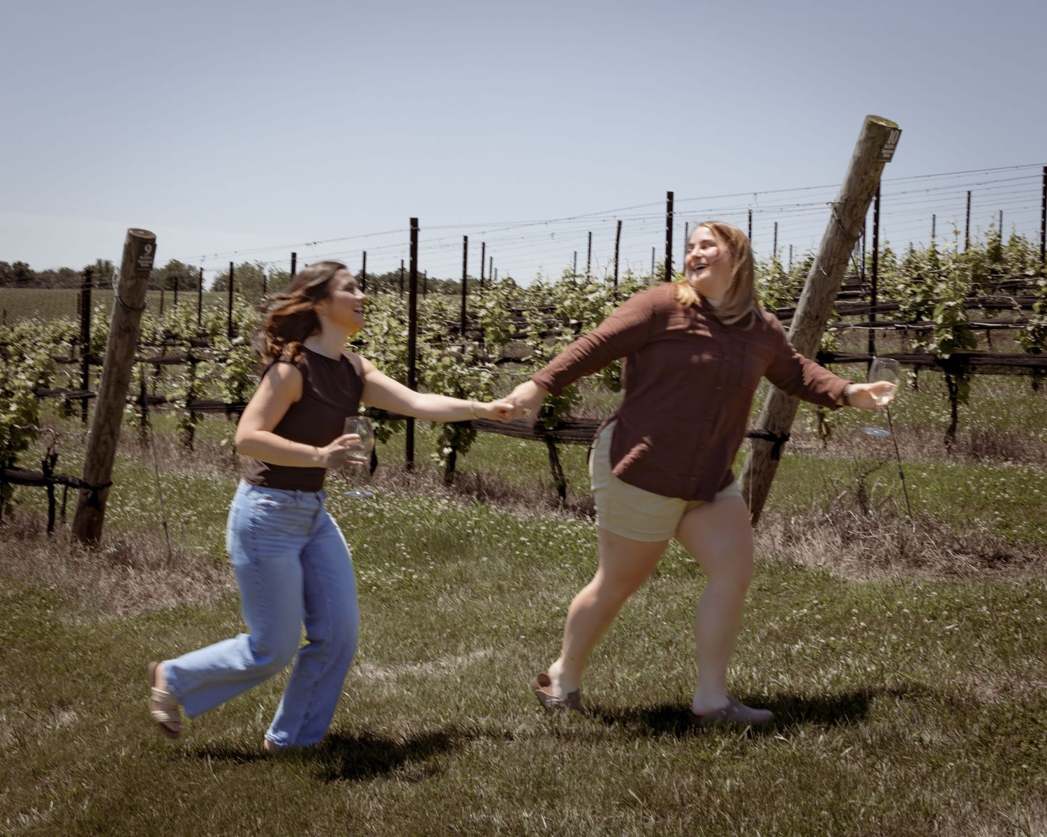 Two women joyfully dancing and holding hands in a sunny vineyard with rows of grapevines in the background.