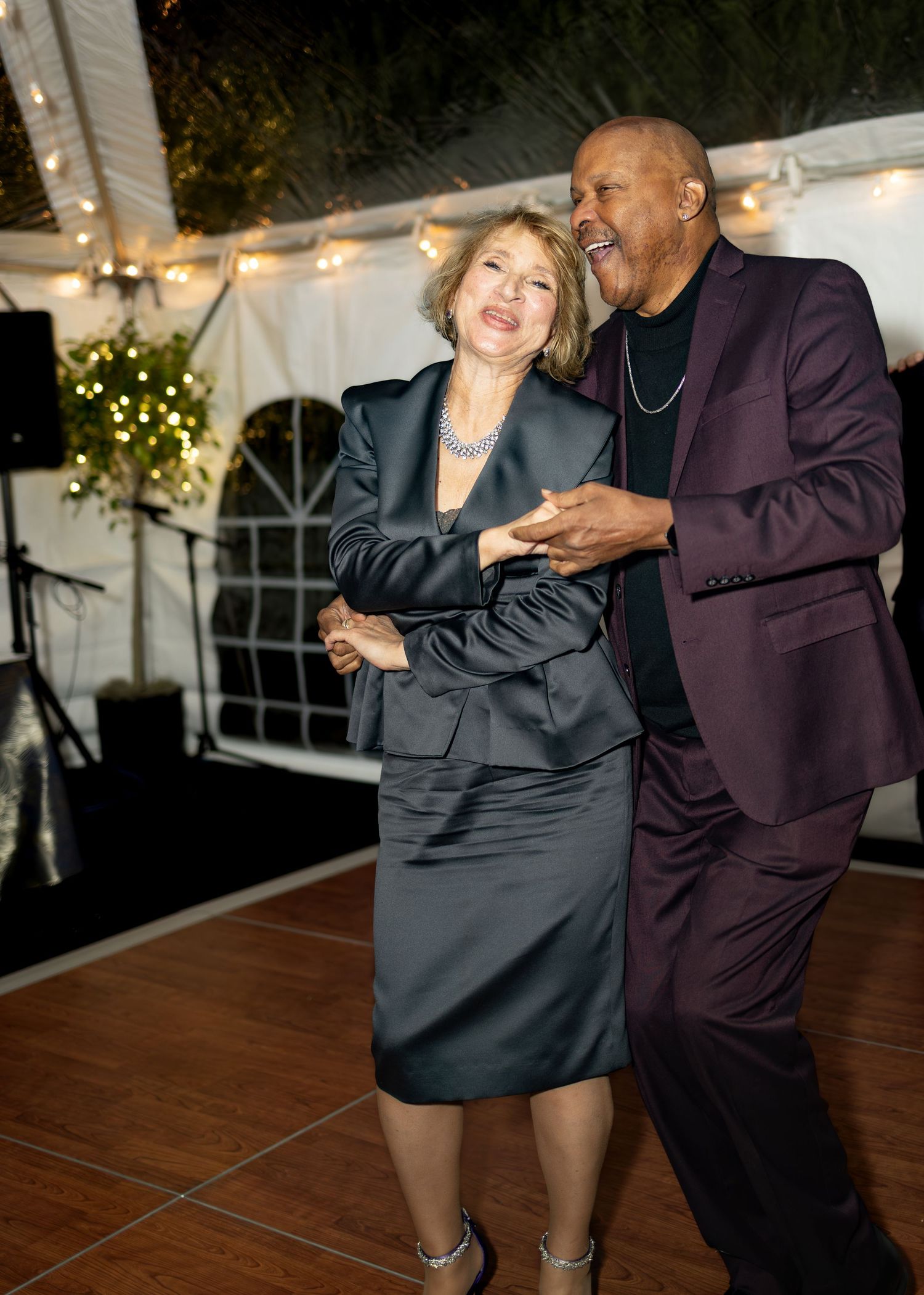 Elegant couple dancing together at a formal event under a white tent with festive string lights.