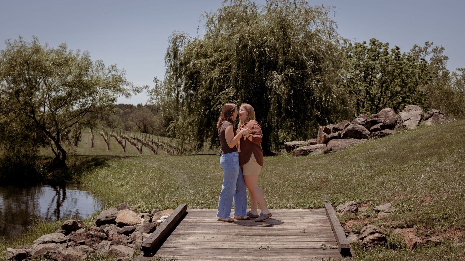 Lesbian couple embracing on a wooden dock by a pond, surrounded by lush green trees and vineyard rows.